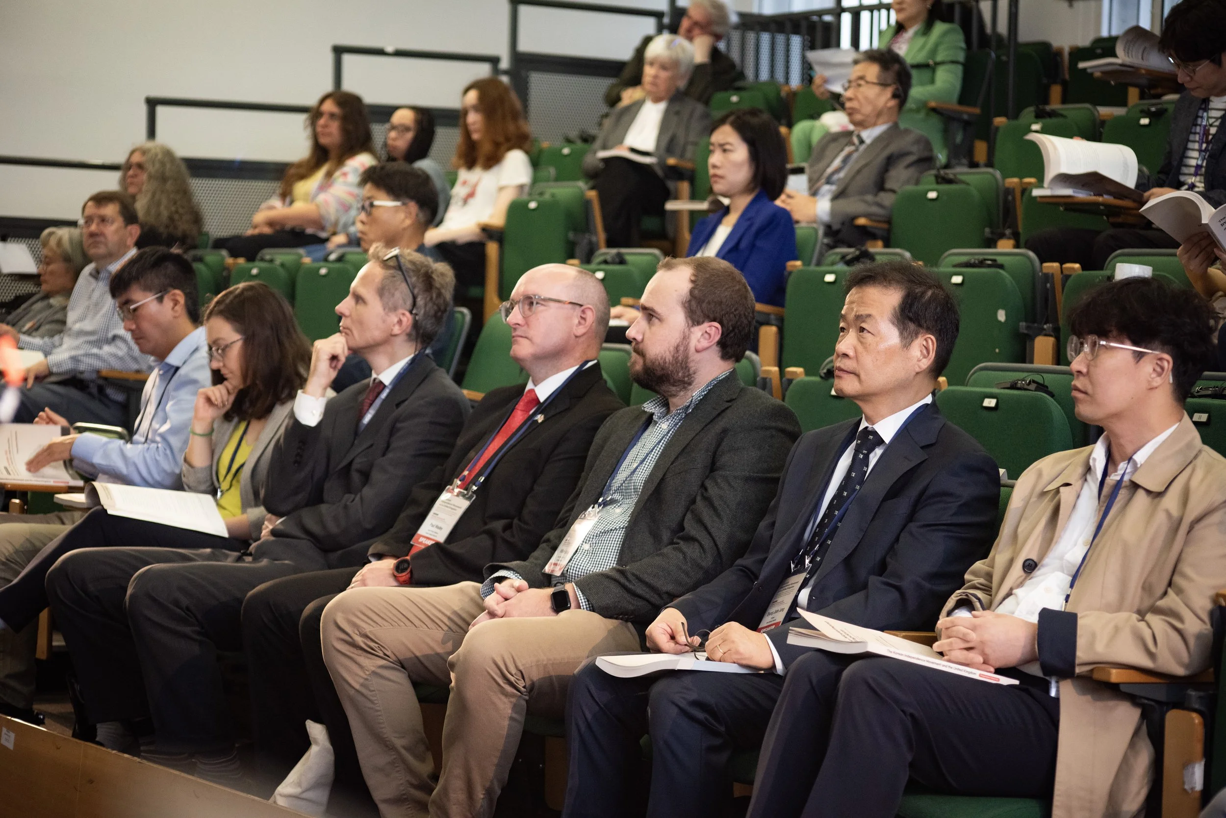 Attendees sitting in green theater seats at a conference, listening to a speaker. Some are taking notes or reading documents.