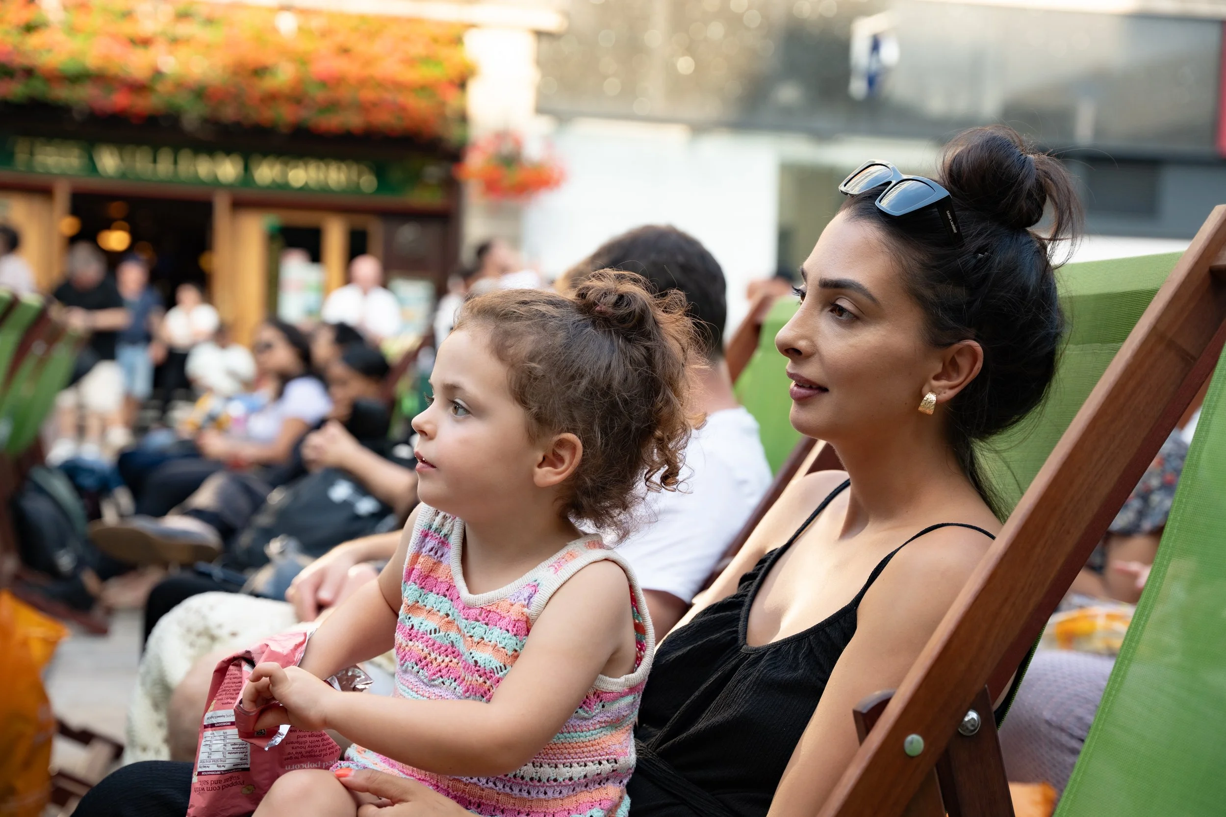 A woman with sunglasses on her head and gold earrings sitting outdoors in a green deck chair with a young girl on her lap during a daytime event, with other people seated behind them