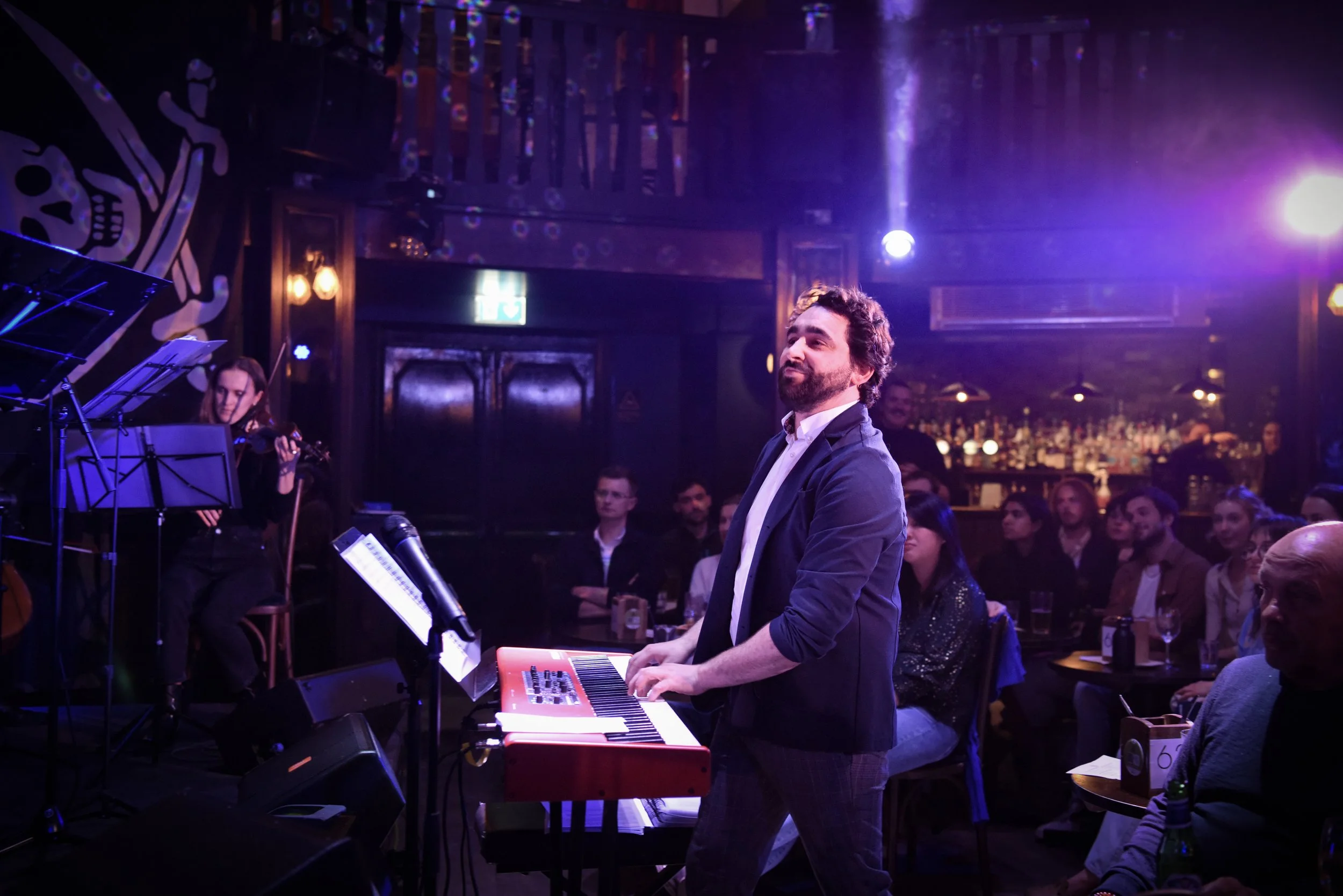 Performer playing keyboard on stage in front of audience in a dimly lit nightclub or concert venue with purple and blue lights.