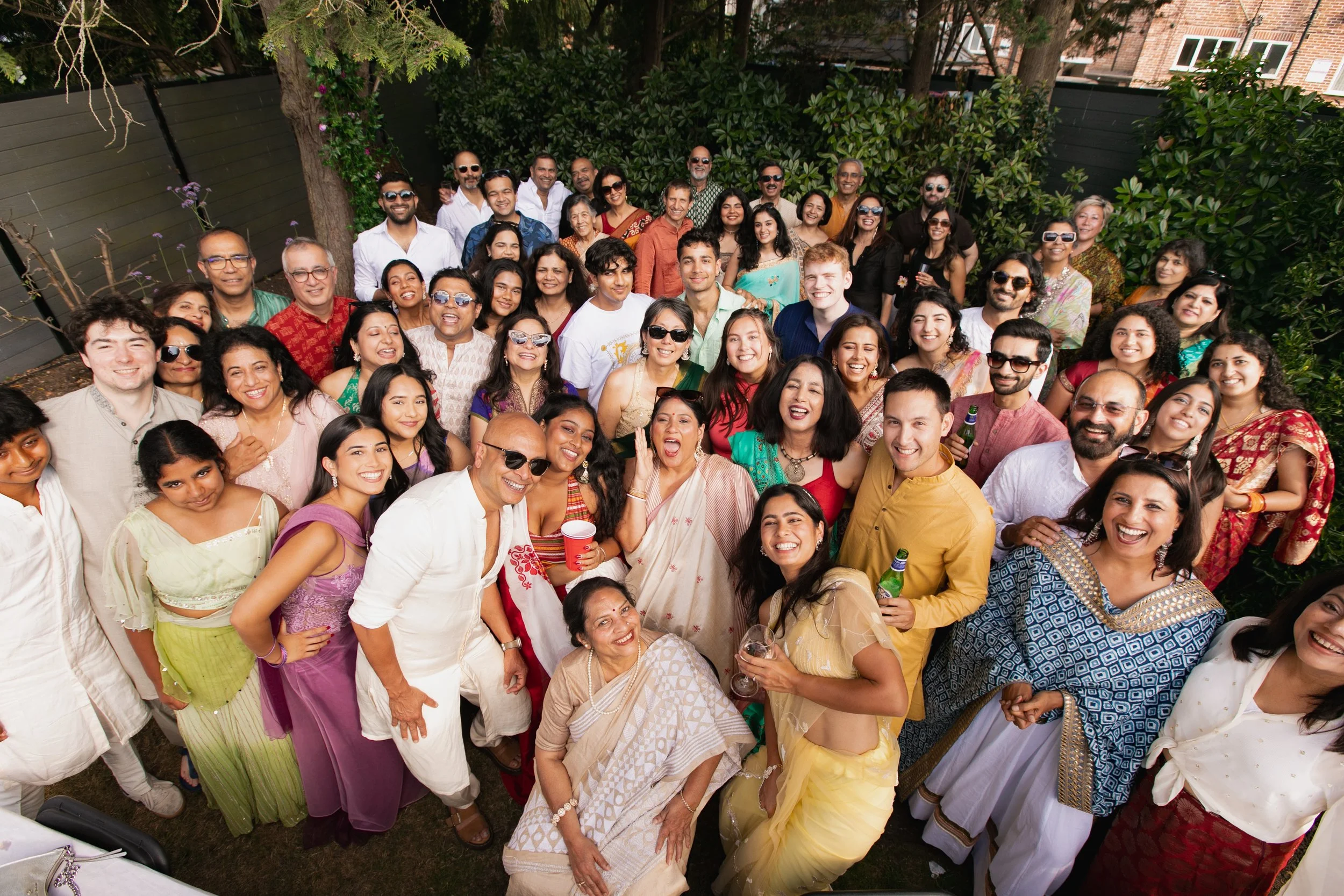 A large group of people gathered outdoors, smiling, and celebrating at a party or wedding, dressed in colorful traditional and modern attire.