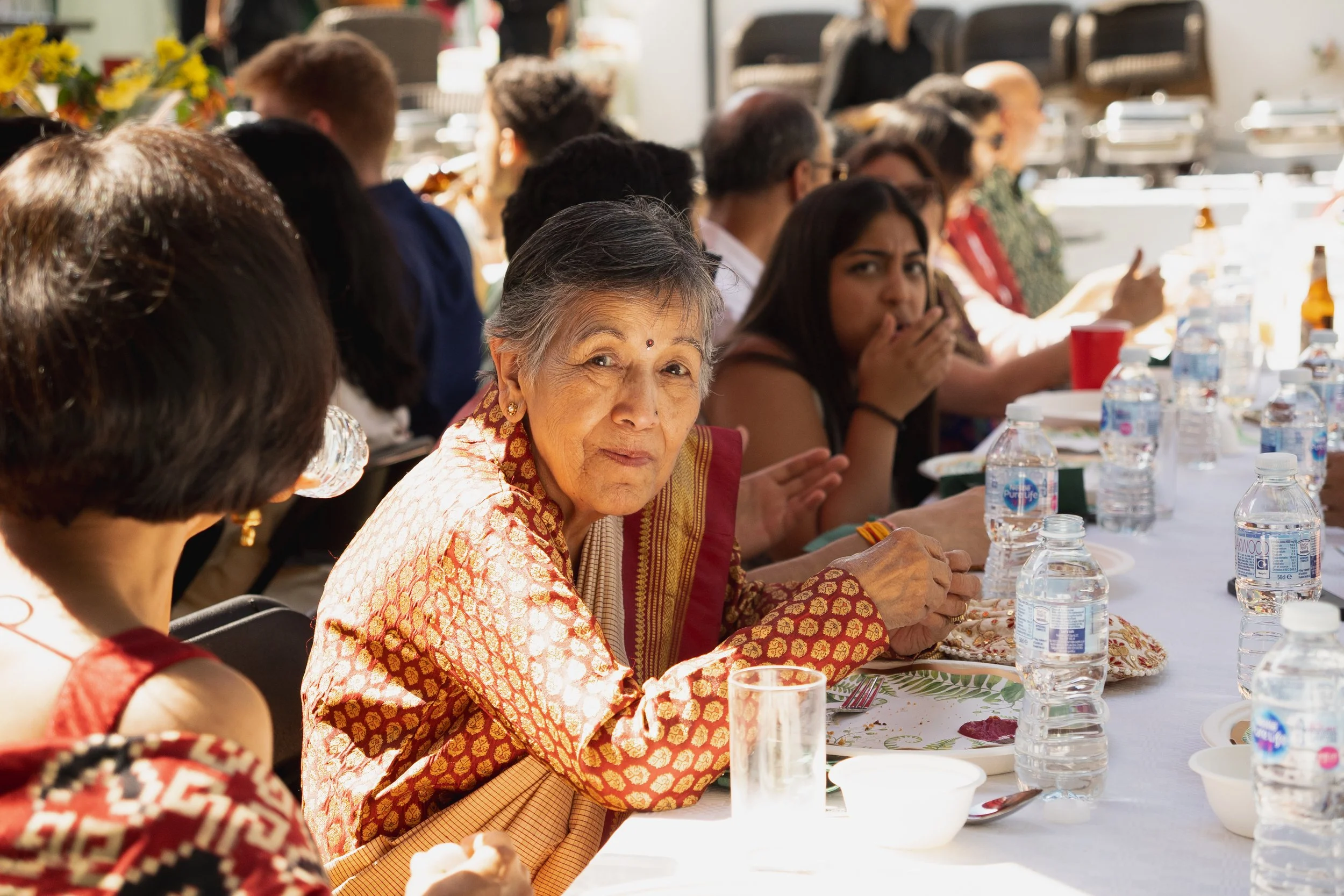 An elderly woman in traditional Indian attire looks at the camera while seated at a long dining table with other people, some of whom are eating or drinking. The table has water bottles, bowls, and plates.