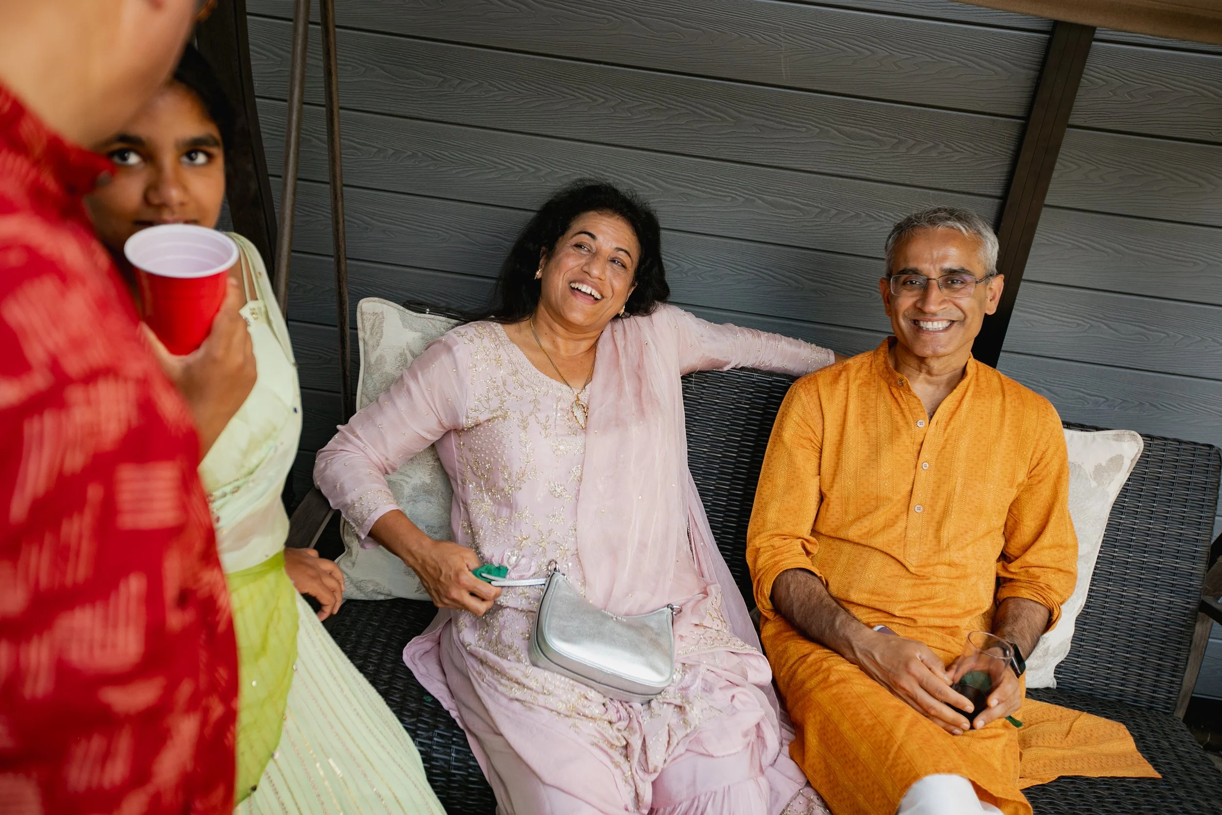 Indian family celebrating together, with two women and one man sitting on a couch, smiling and engaging in conversation, dressed in traditional attire.