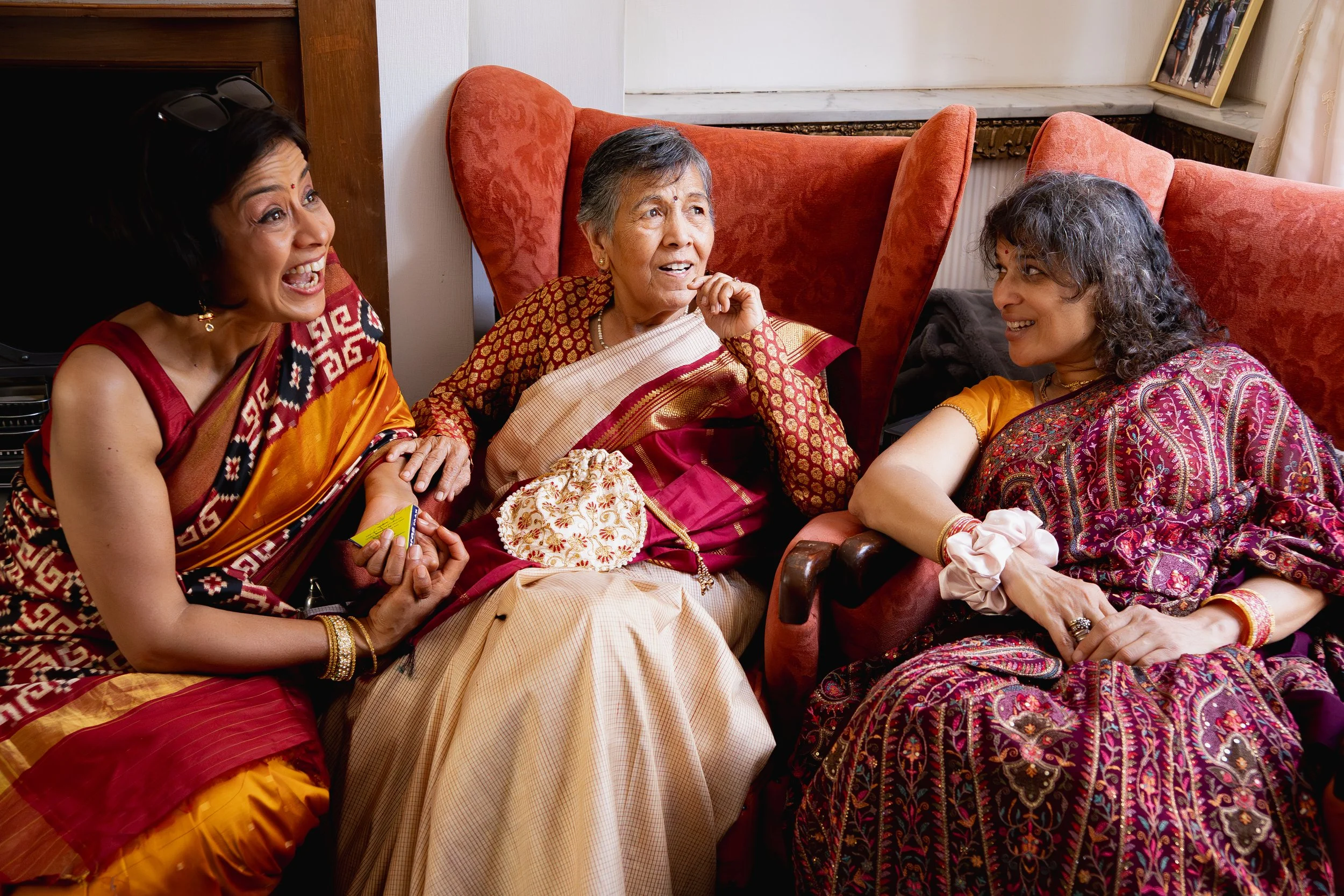 Three women wearing colorful traditional Indian sarees, sitting and smiling in a cozy room with red armchairs and a framed photo on the wall.