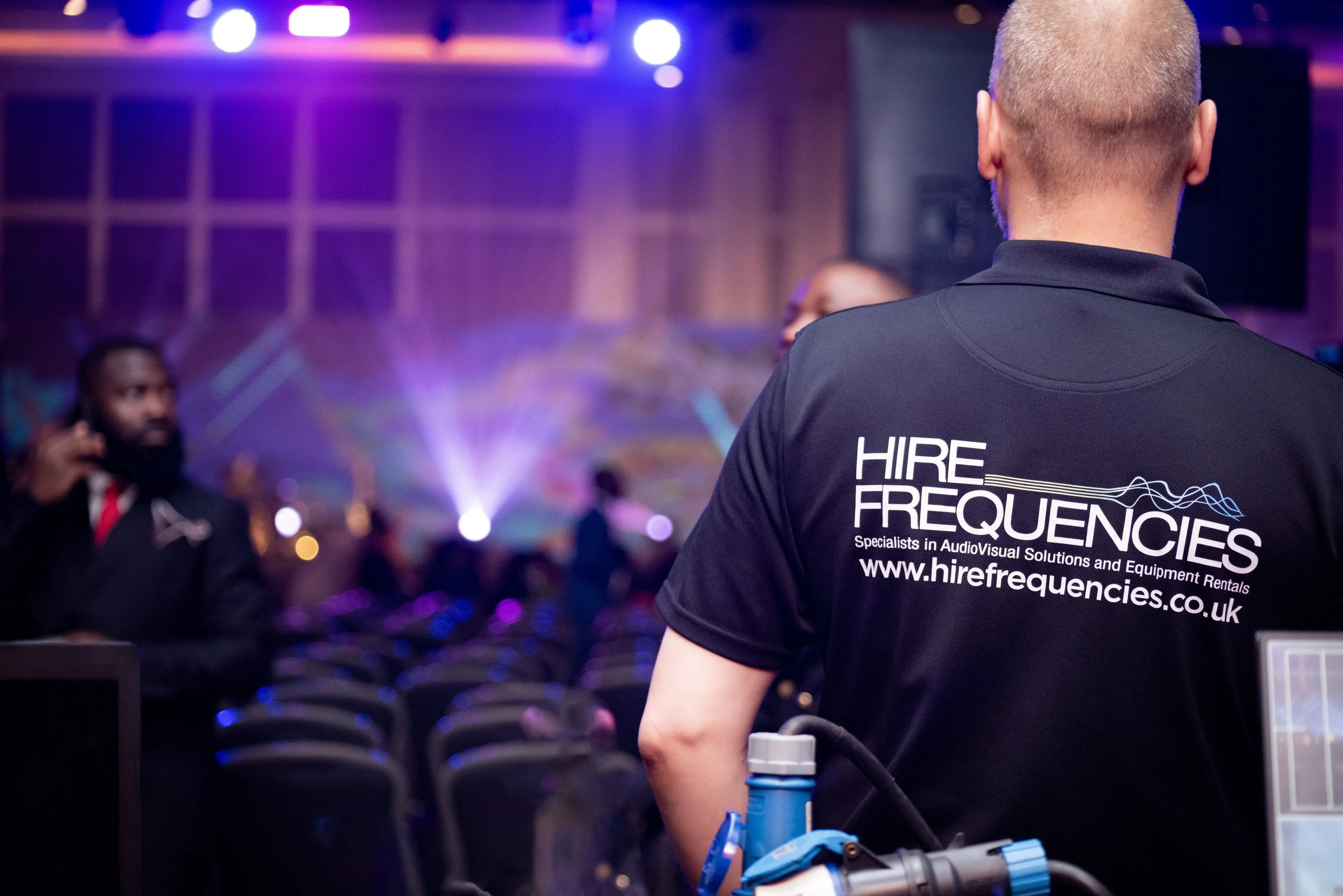 Event staff wearing a black shirt with 'HIRE FREQUENCIES' logo seen at a vibrant indoor conference or concert venue with colorful stage lights and audience seating.