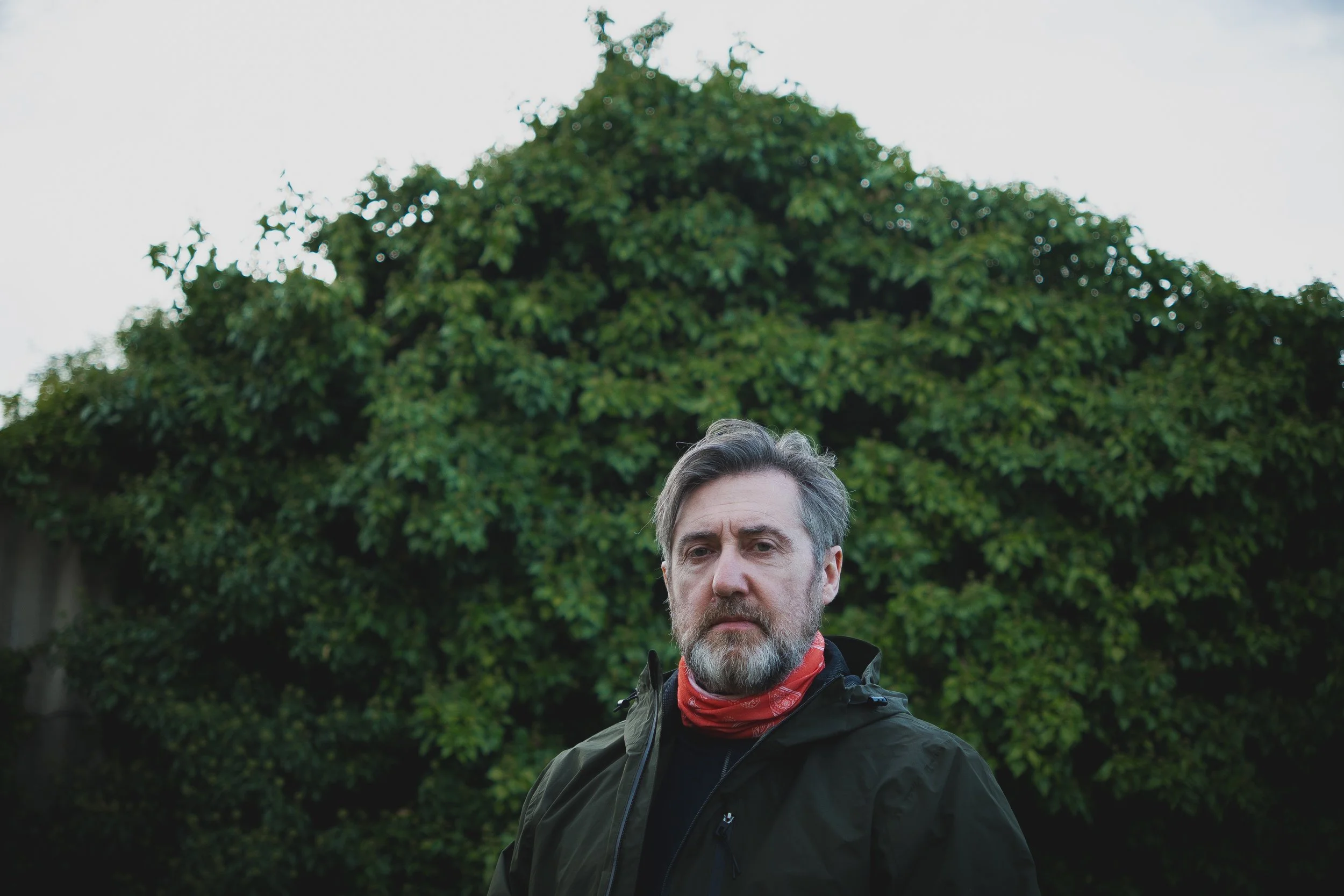 A middle-aged man with a beard and gray hair, wearing a dark jacket and a red bandana around his neck, standing outdoors in front of a large leafy green bush or tree.