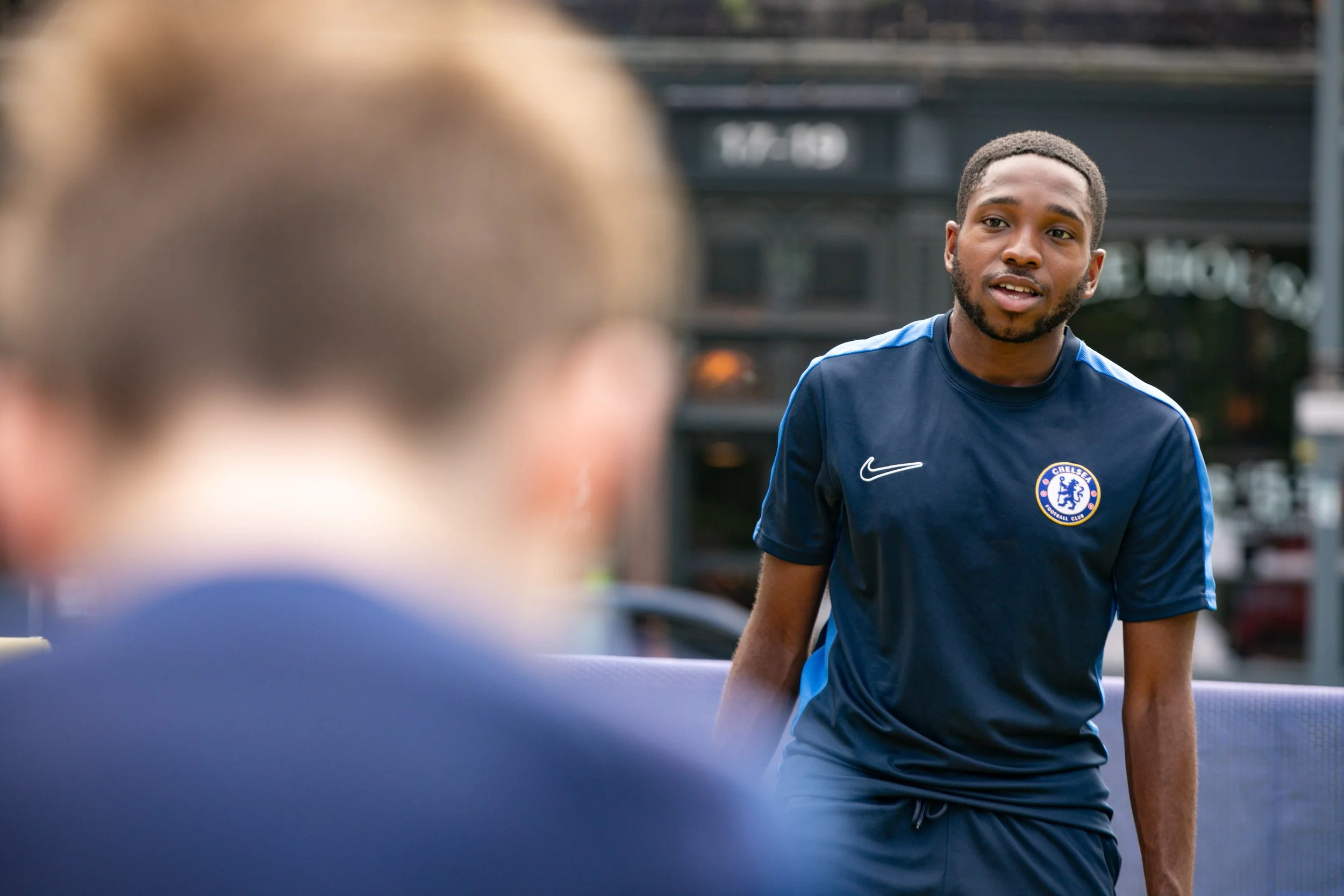A young man in a navy blue Chelsea football training kit, standing outdoors, speaking to a person in the foreground who is out of focus.