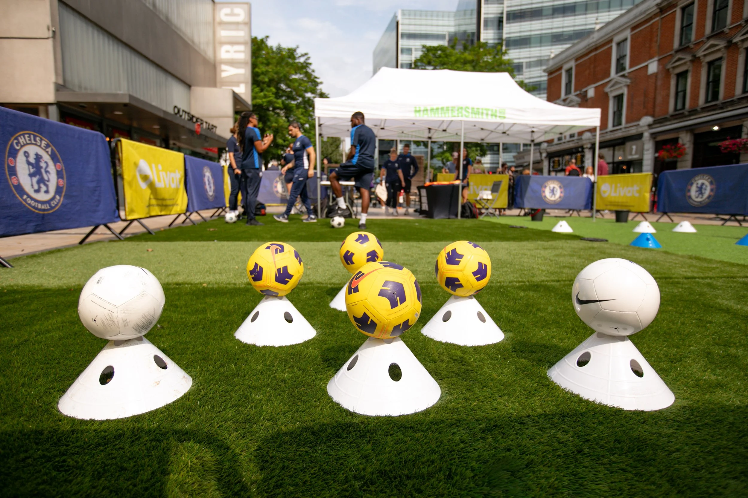 Soccer training setup with foam cones and soccer balls on artificial grass, in an outdoor urban area with a Chelsea Football Club banner, people practicing in the background, and a white tent for the event.