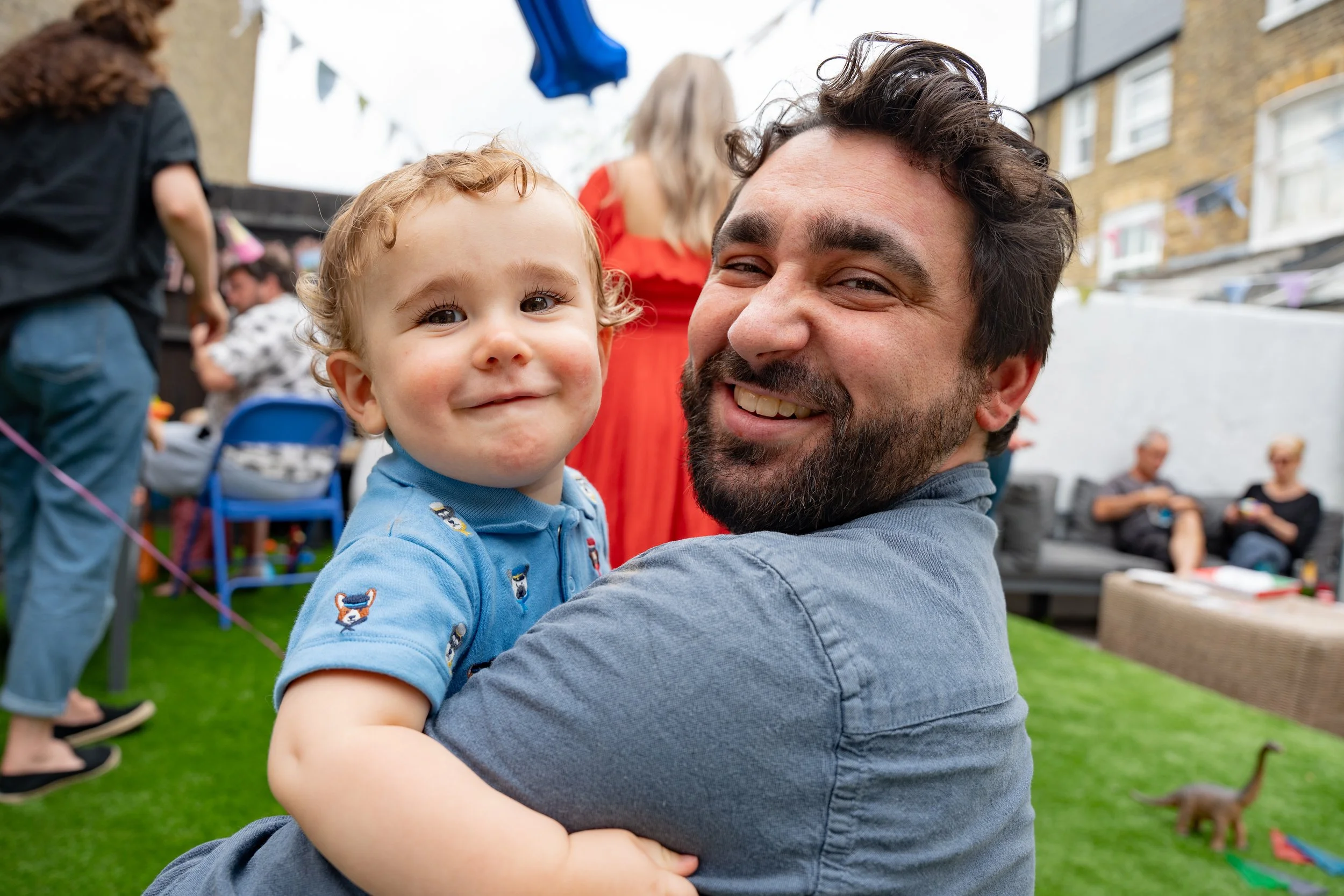A man holding a young child, both smiling, at an outdoor party with other people and toys in the background.