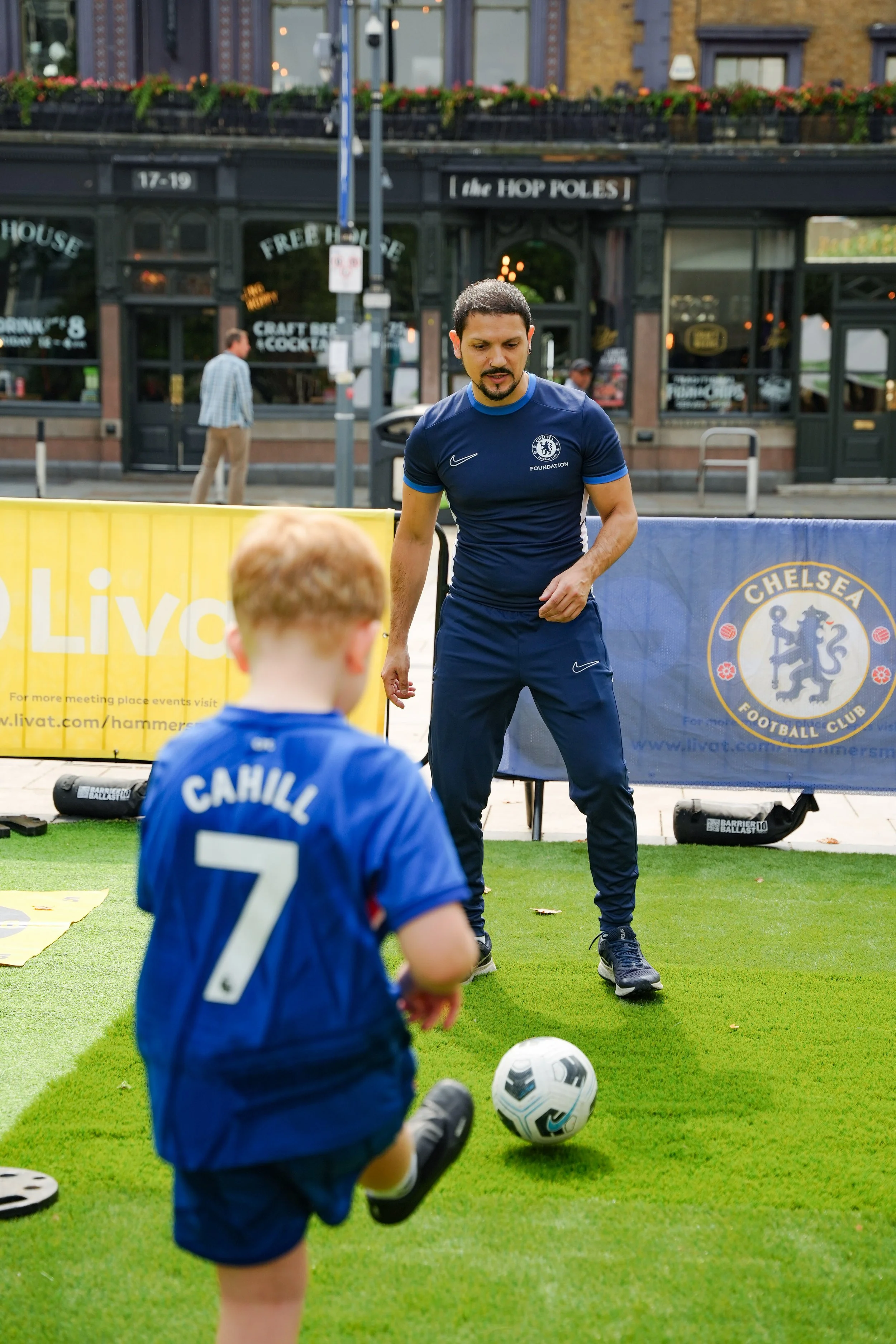 A man in a Chelsea football club shirt instructs a young boy in a blue jersey, numbered 7 and named Cahill, during a soccer training session on a green field. A Chelsea FC logo banner is visible in the background.