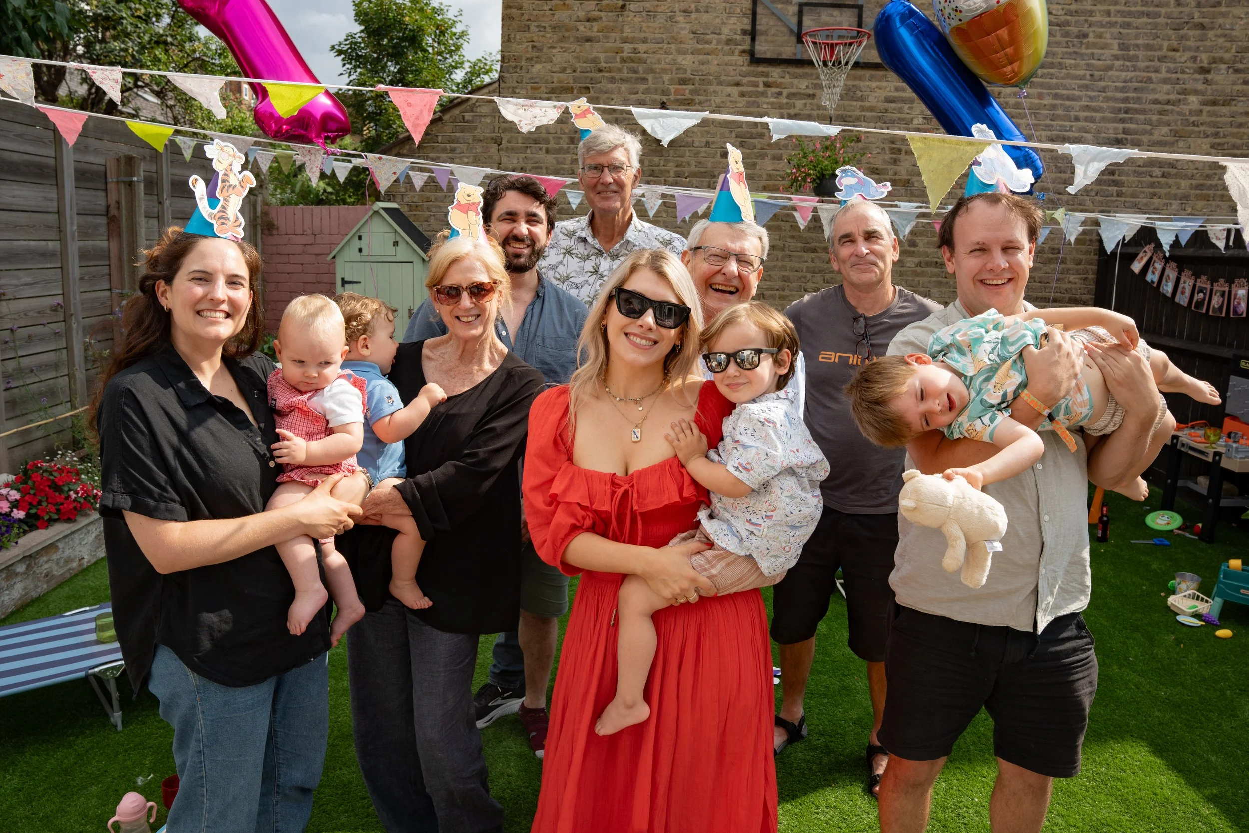A group of people celebrating outdoors with party decorations, children, and adults smiling, some wearing birthday hats.
