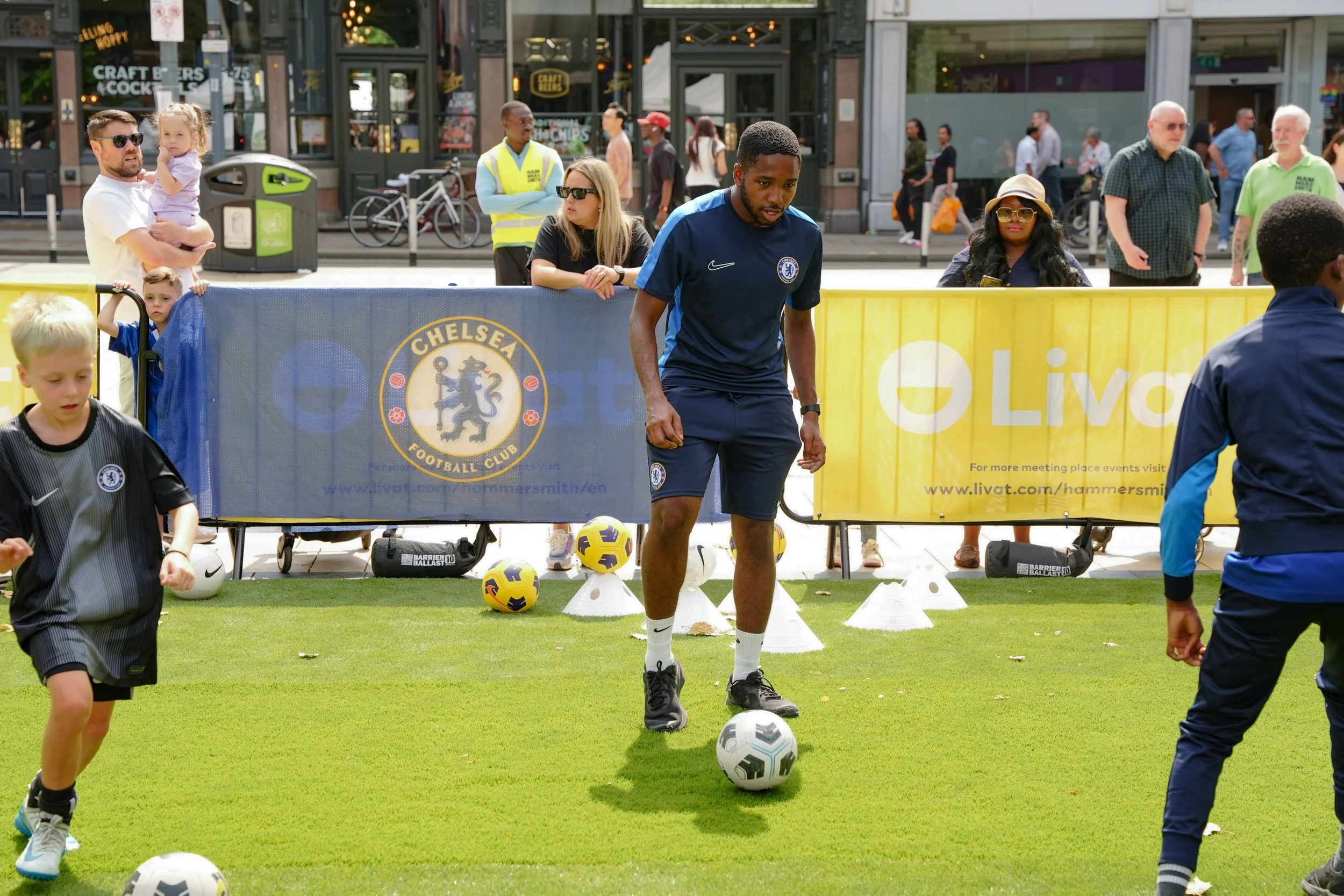 Soccer coach practicing ball skills with children on a field in an urban setting, with spectators and city street in the background.
