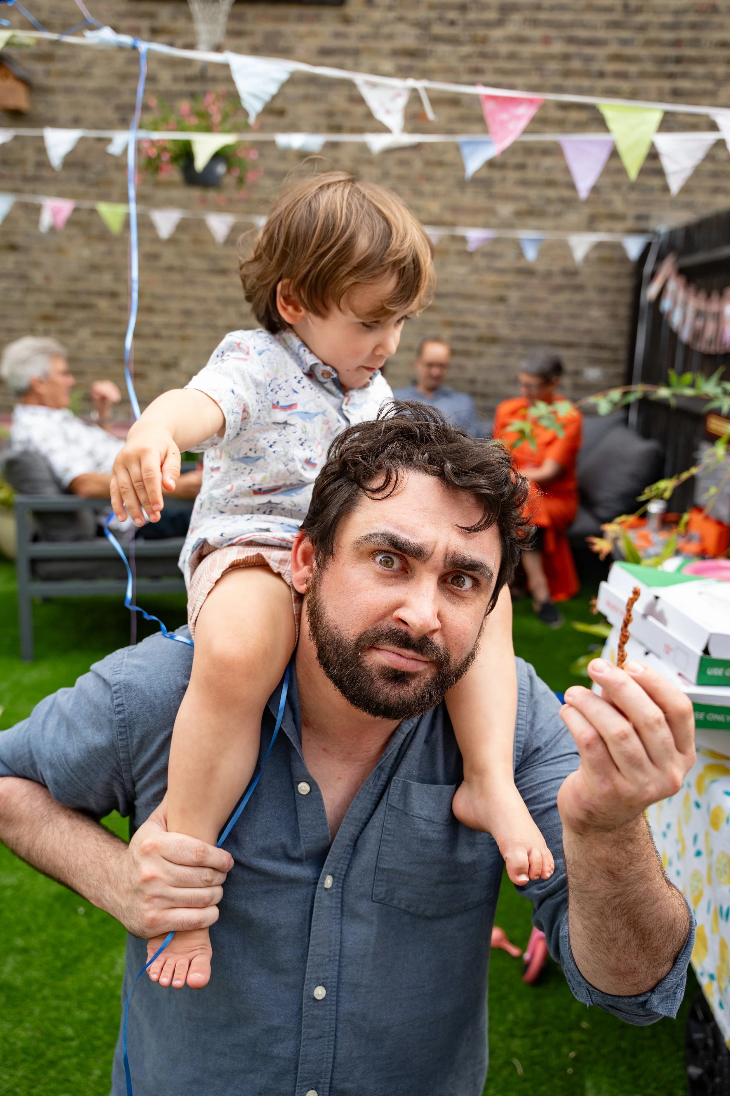 A man with dark hair and a beard carrying a young boy on his shoulders at an outdoor party. The boy is reaching for a small birthday cake or cupcake with a curious expression. Bunting flags and other party decorations are visible in the background, w