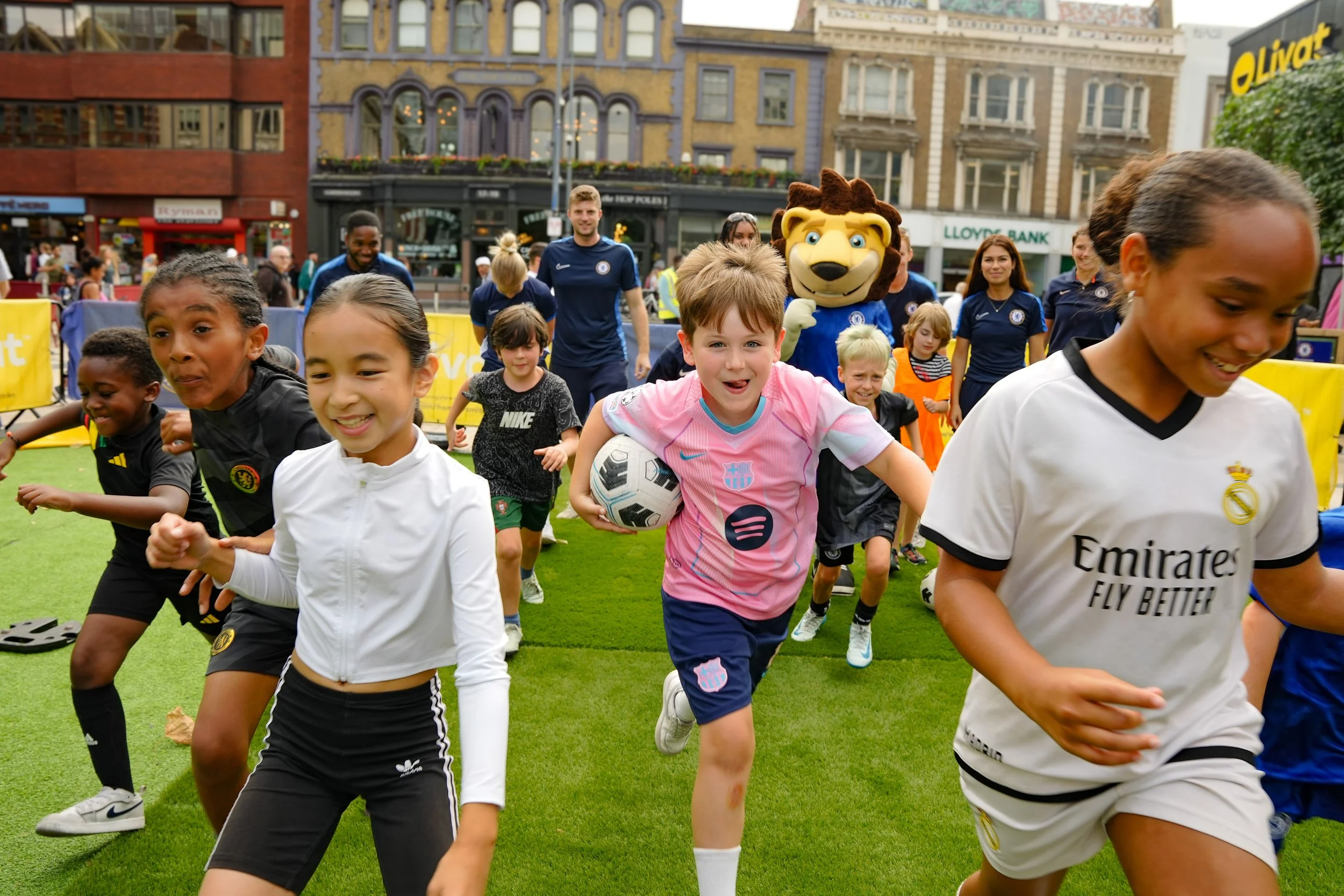 Children running on a green field during a soccer event with a lion mascot and adults in the background.