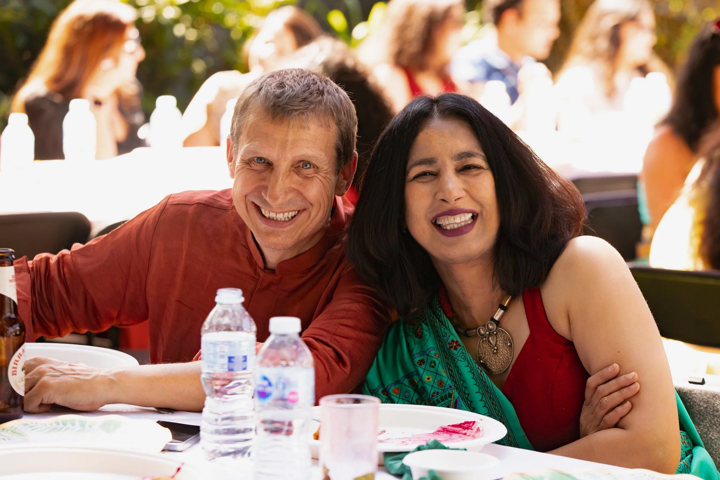 Smiling man and woman sitting at a table outdoors during a celebration or gathering, with other people in the background.