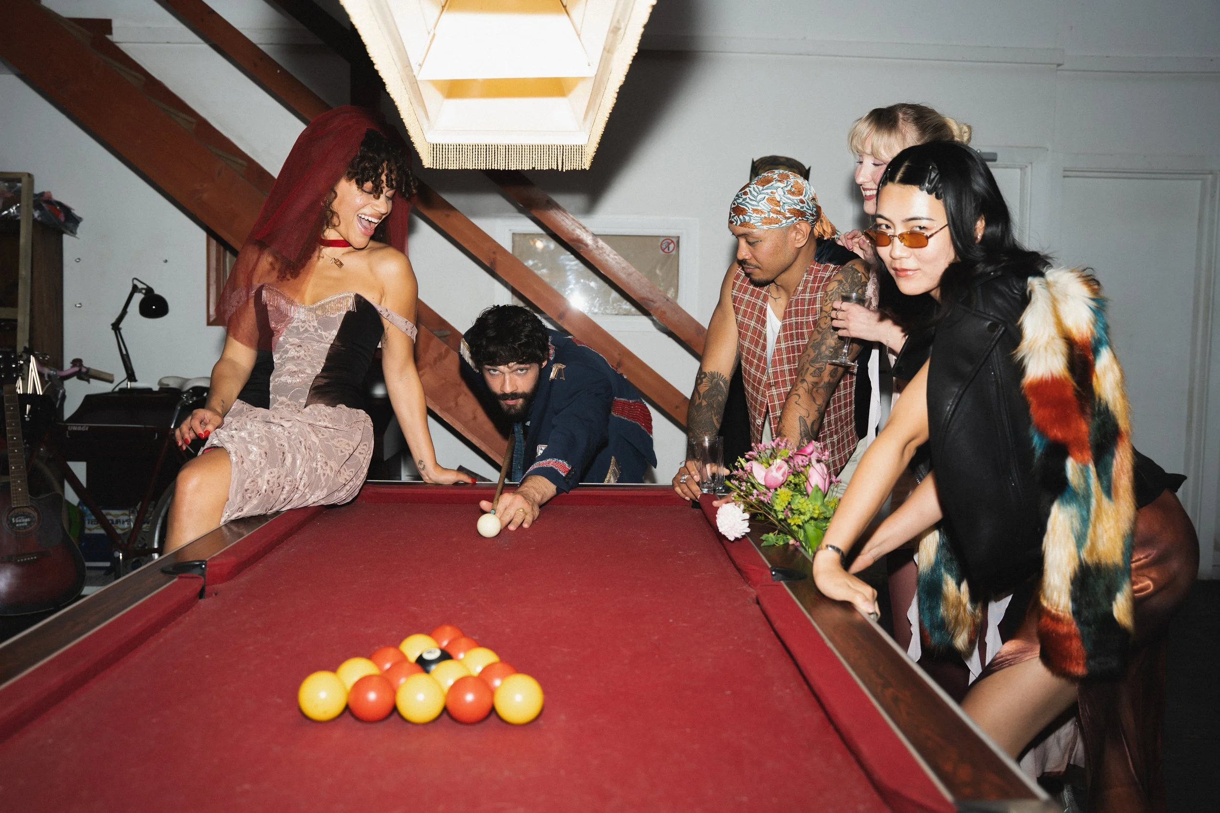 Group of five friends playing billiards, with one woman leaning on the table, smiling, and four others watching and smiling, in a cozy indoor setting with musical instruments in the background.