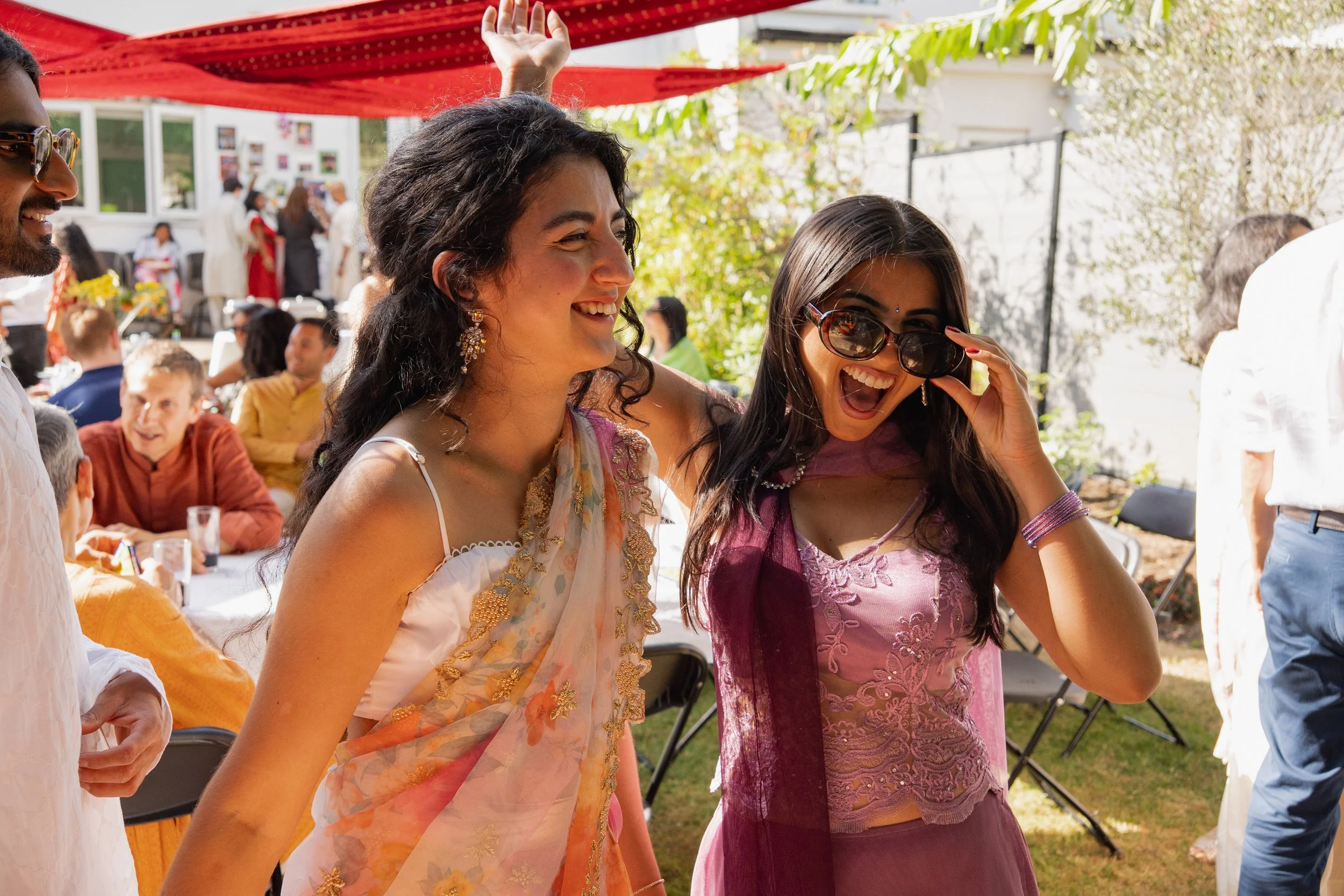Two women celebrating together at an outdoor event, smiling and laughing, one adjusting sunglasses. Other people are in the background, seated and standing at tables, with festive decorations and greenery.