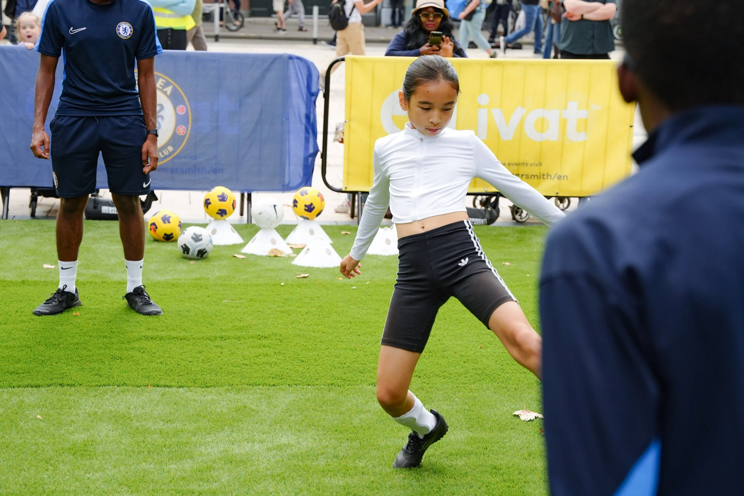 A young girl is playing or practicing soccer on a grassy field, surrounded by a small group of people and soccer balls.