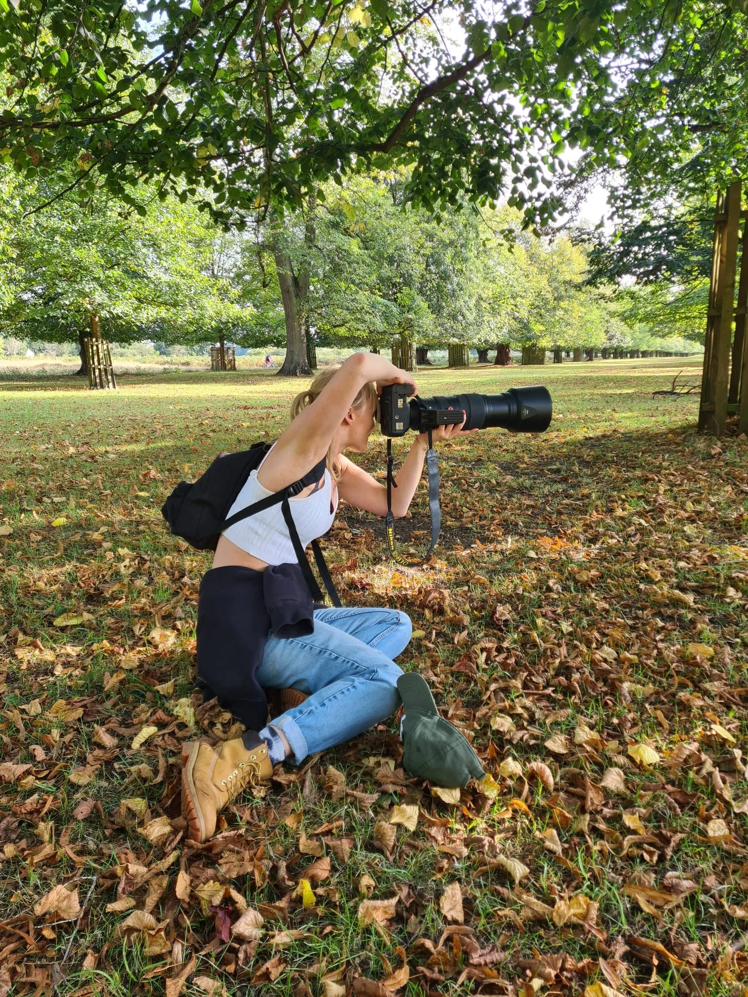A woman kneeling on the ground, taking a photograph with a camera and telescope in a park with trees and fallen leaves.