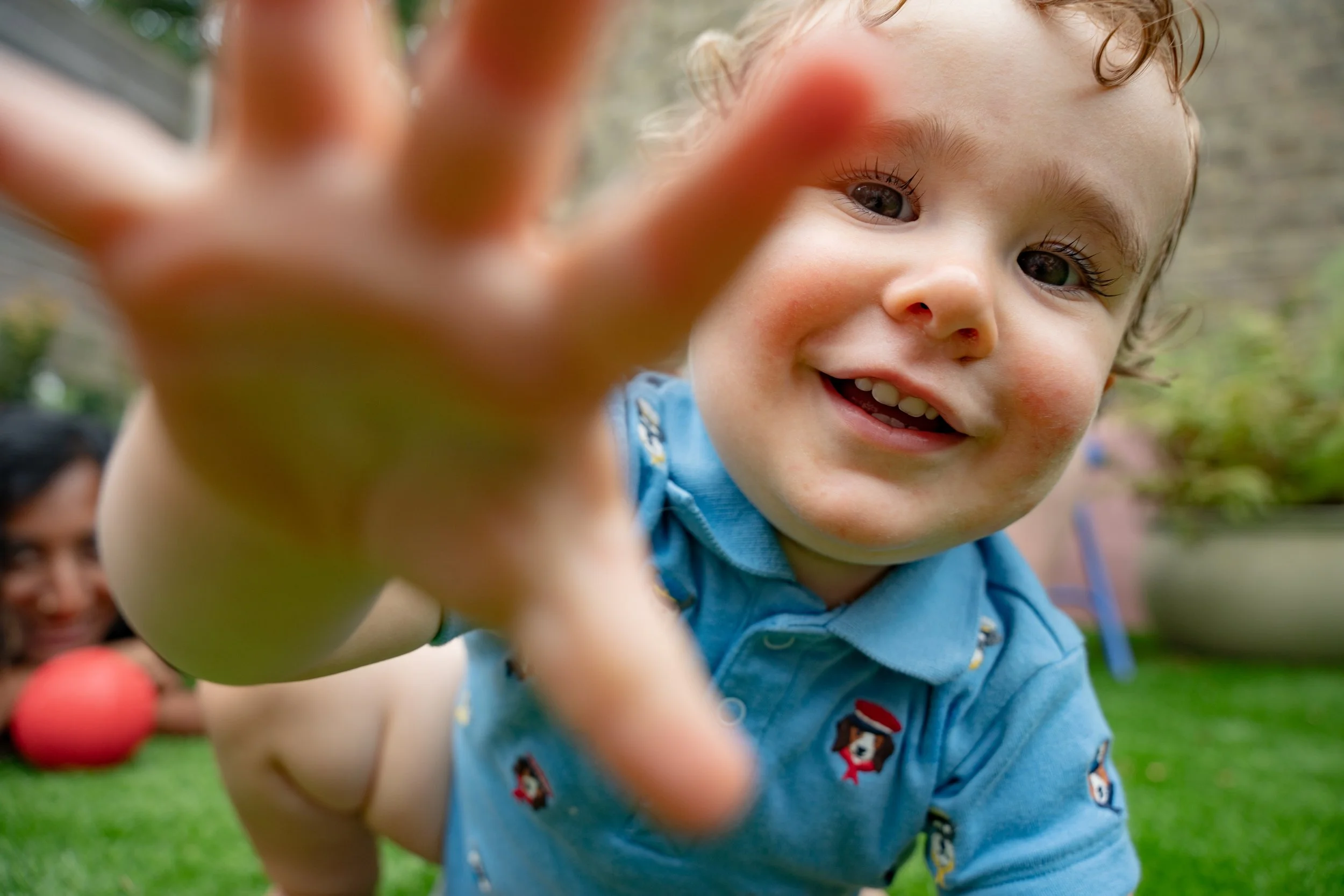 Close-up of a smiling young boy reaching toward the camera outdoors, with a woman and a garden in the background.