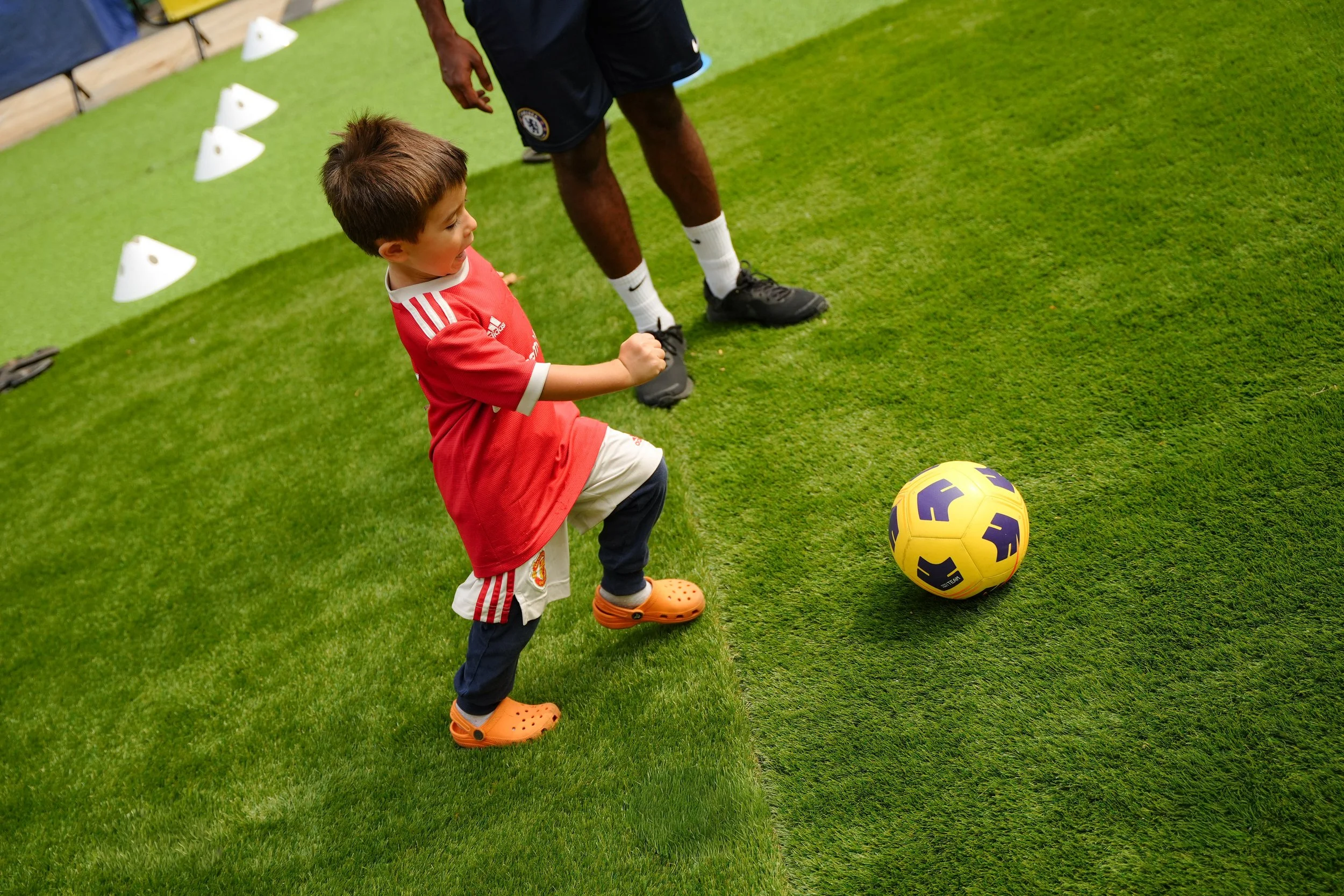 A young boy playing soccer on a green field with a soccer ball nearby, while an adult watches. The boy is wearing an orange Croc shoe, a red sports jersey, and soccer shorts.