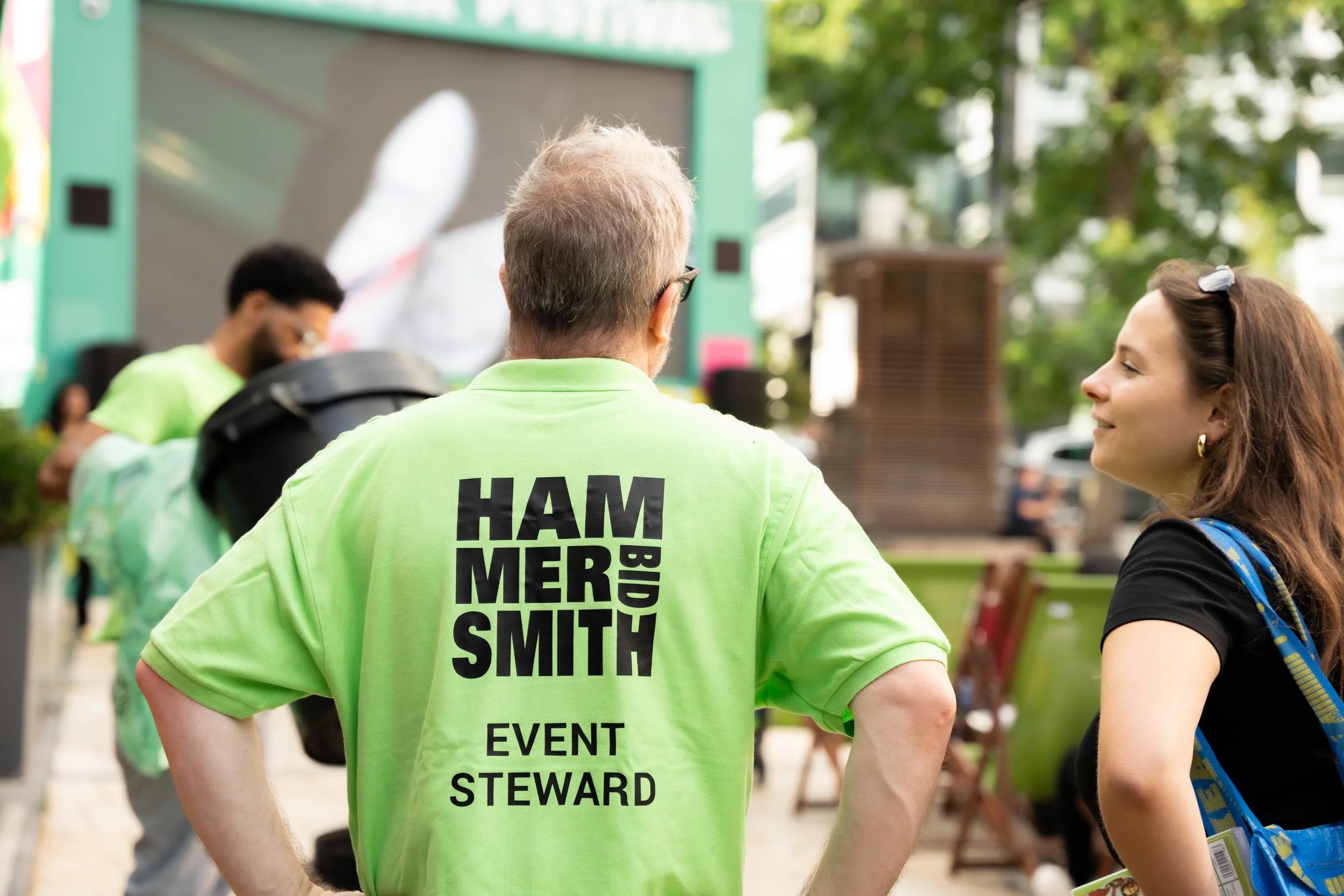 A man in a bright green event staff shirt talking to a woman at an outdoor event.