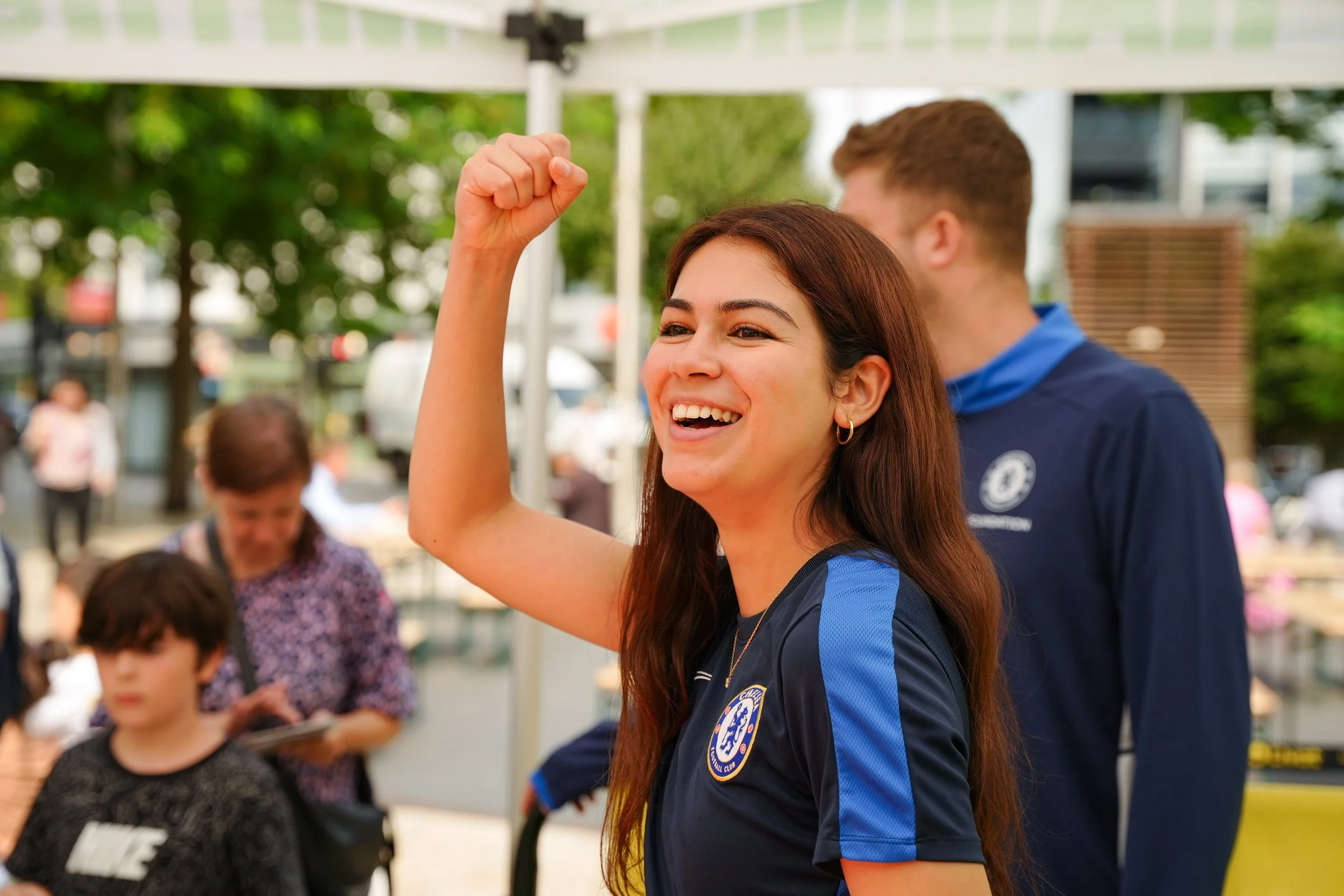 A young woman wearing a Chelsea football club jersey is smiling and raising her fist in celebration. She has long brown hair and small hoop earrings. A man standing behind her is also in a Chelsea jersey. The background shows other people and a park 