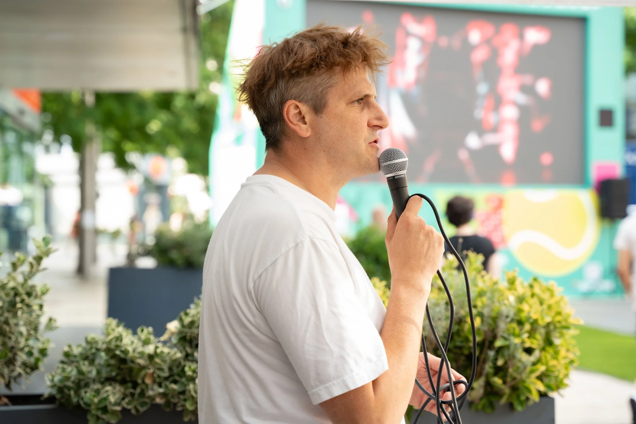 A man with short, tousled hair in a white t-shirt holding a microphone at an outdoor event.