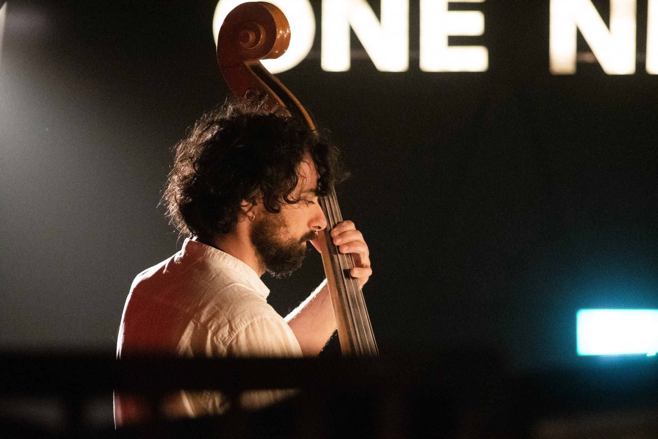A man with curly dark hair and a beard playing a double bass on stage at a concert, with a large illuminated sign with partially visible text in the background.