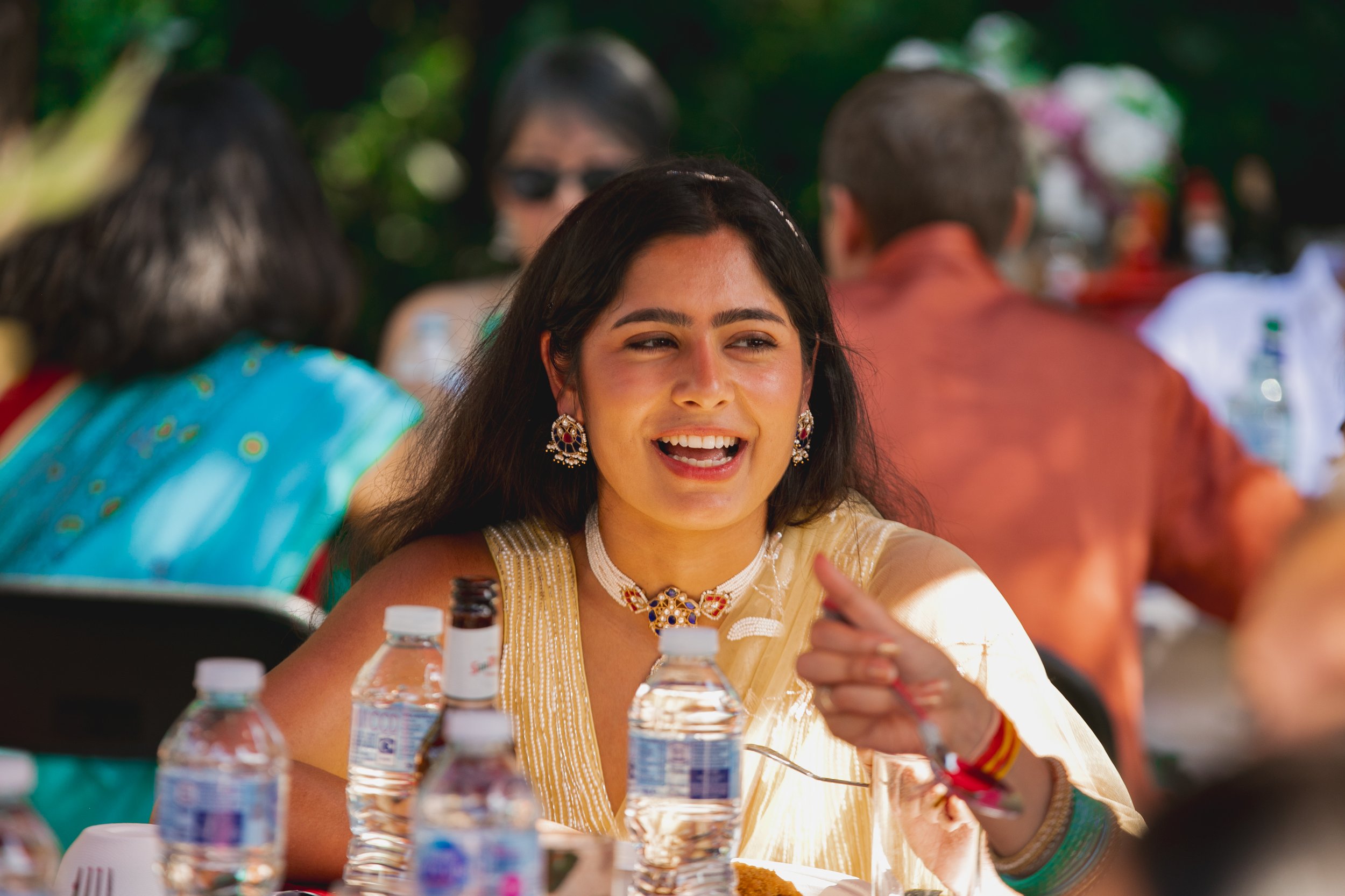 Young woman smiling and talking at a social outdoor gathering, wearing traditional attire and jewelry, with water bottles and food on the table.