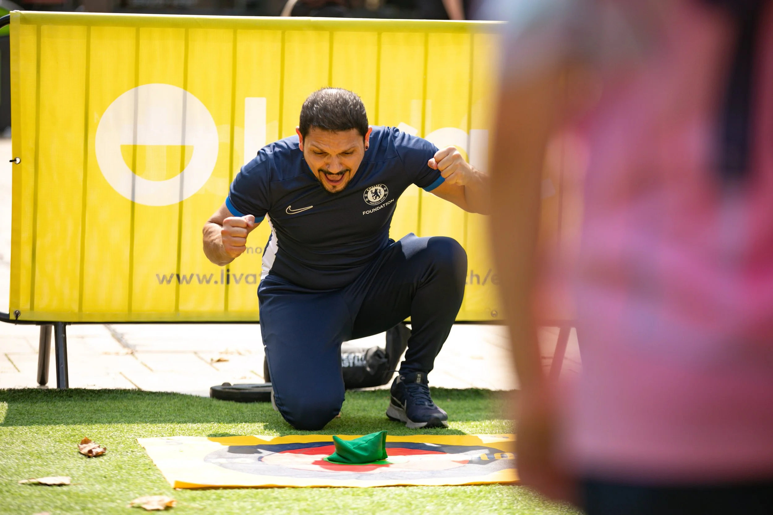 A man kneeling on the ground cheering with clenched fists in front of a yellow banner and a green hat on a mat, with a woman in pink partially visible in the foreground.