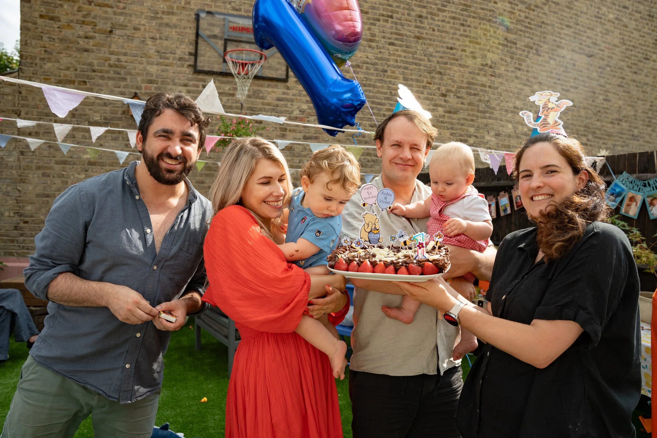 Group of people celebrating a birthday party outdoors, holding a birthday cake with strawberries and decorations, with birthday hats and party decorations in the background.