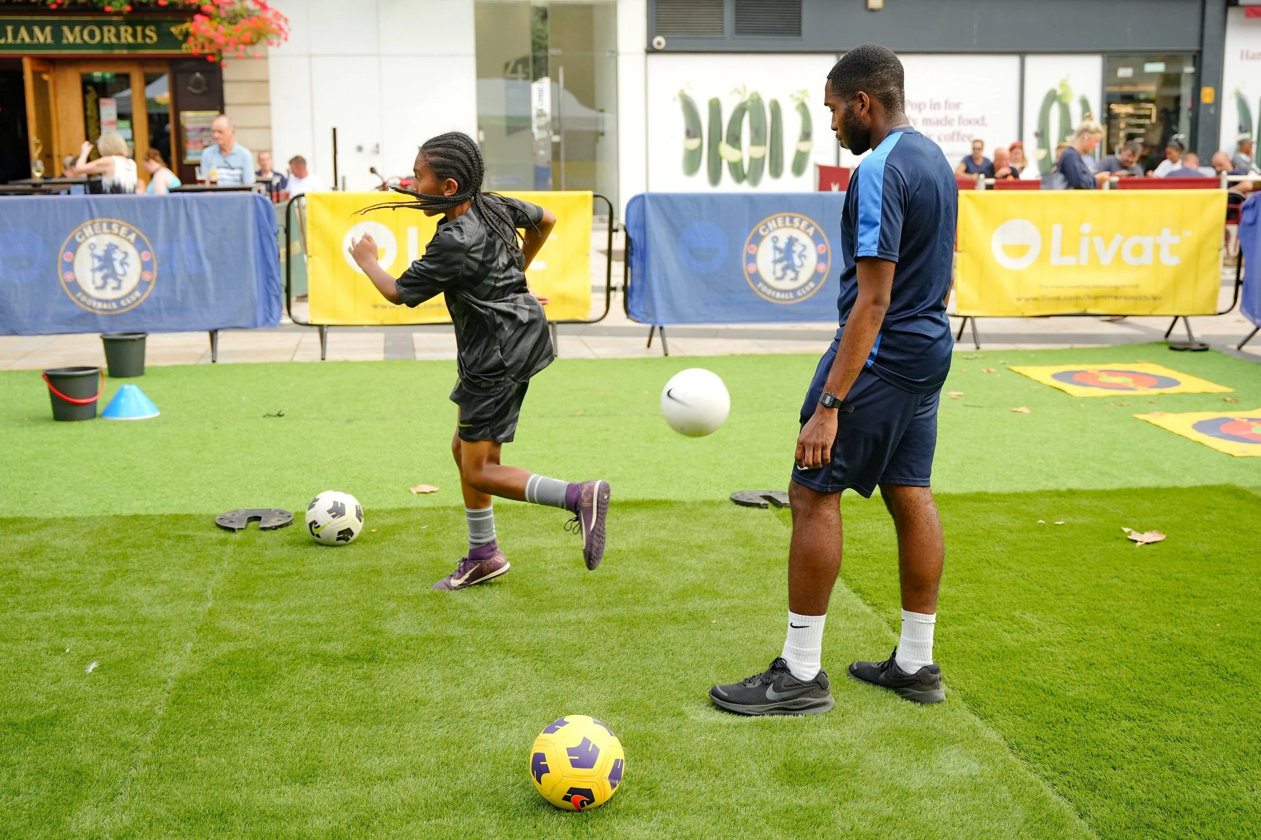 A young girl playing soccer in a public area outdoors, with an adult man observing nearby. The girl is kicking a soccer ball, and there are additional soccer balls and cones on the green artificial turf. People are sitting at tables in the background