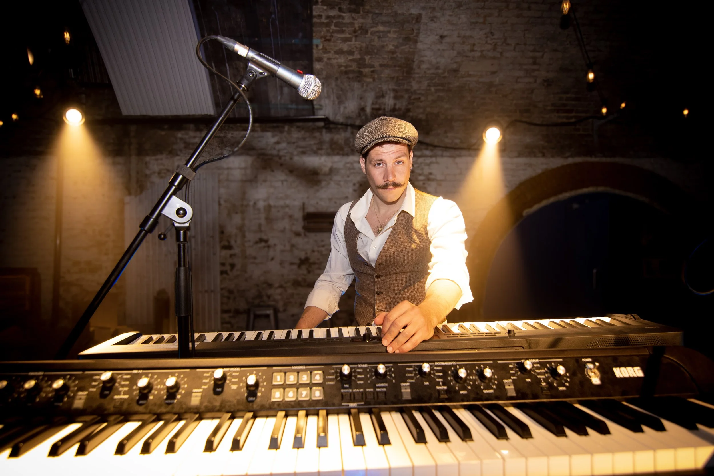 A man with a mustache and wearing a vest, white shirt, and a flat cap playing a keyboard with a microphone nearby in a dimly lit brick-walled venue.