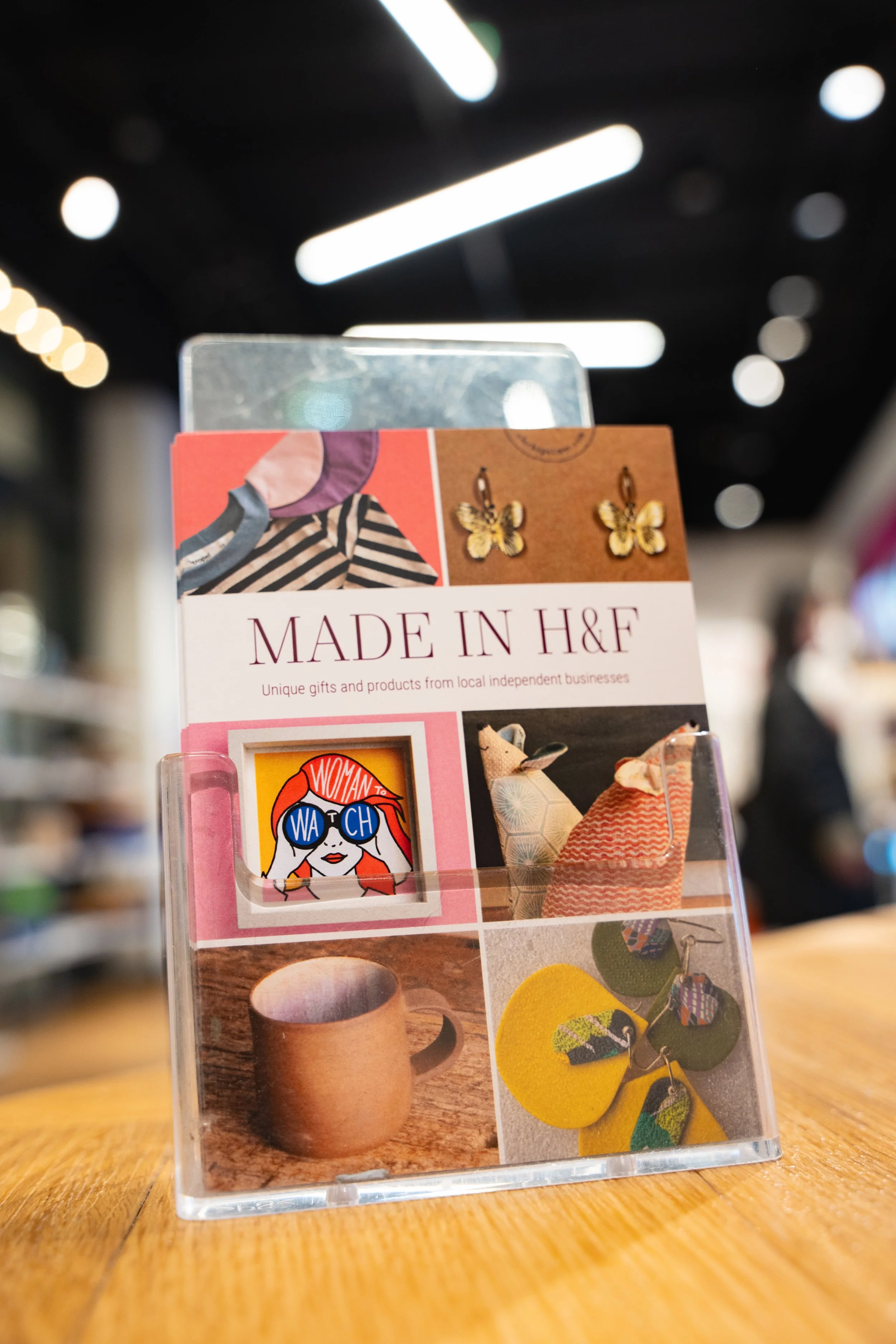 Brochure displaying handmade jewelry, accessories, and home decor items from local independent businesses, with images of butterfly earrings, a colorful women’s graphic art, a ceramic mug, and fabric accessories on a wooden surface.