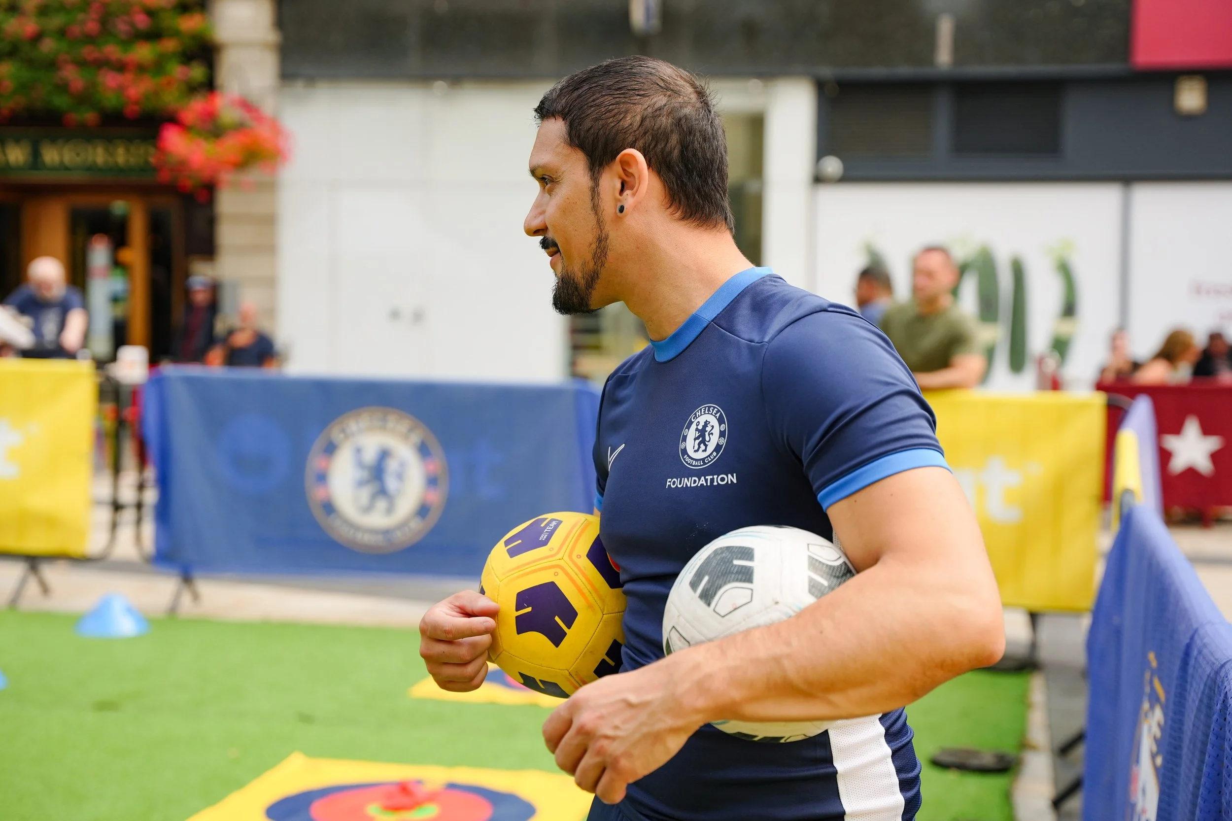A man in a Chelsea football jersey holding multiple soccer balls during an outdoor event.