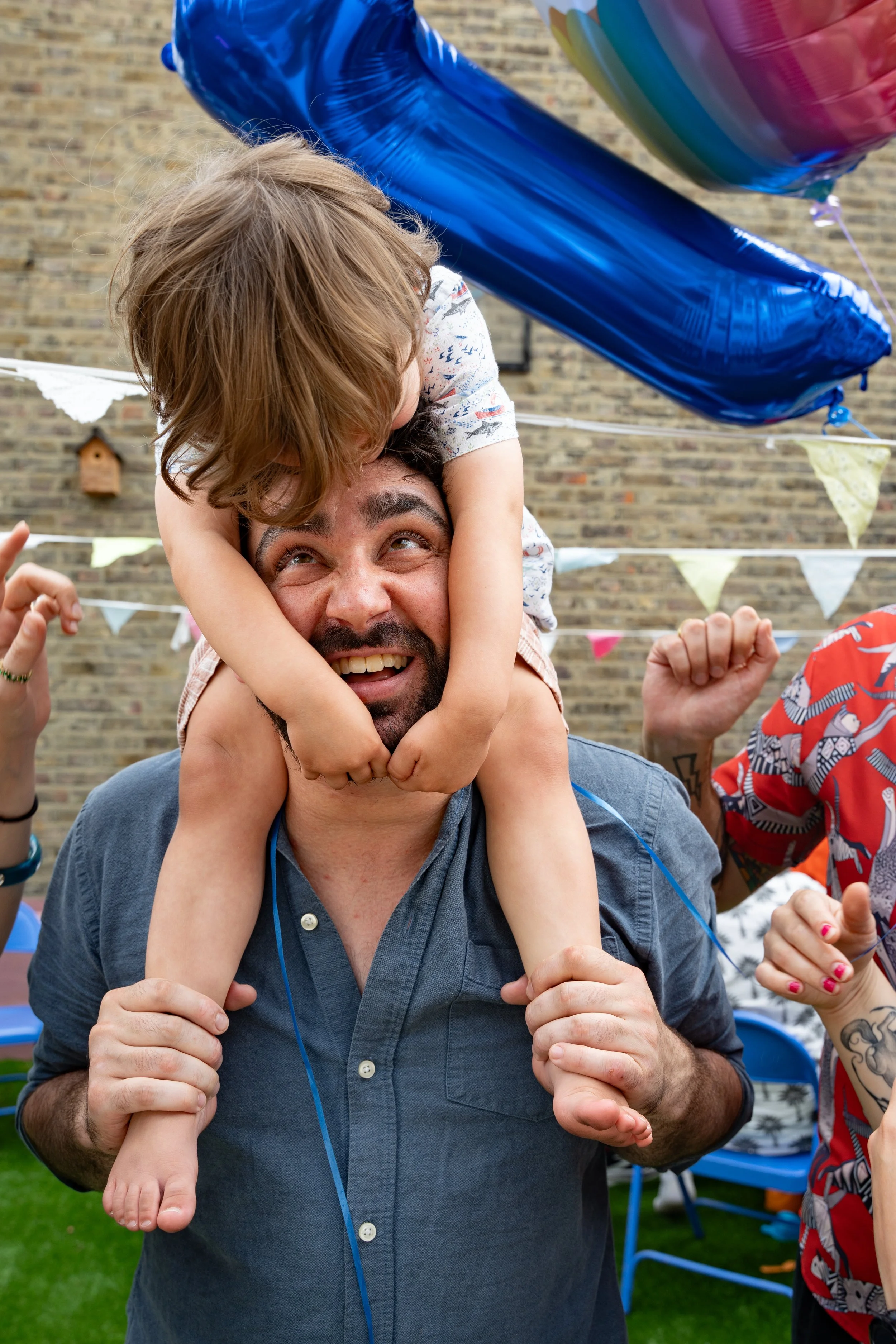 A man with a beard smiling as he carries a young child on his shoulders at an outdoor birthday party, with colorful balloons and bunting in the background.