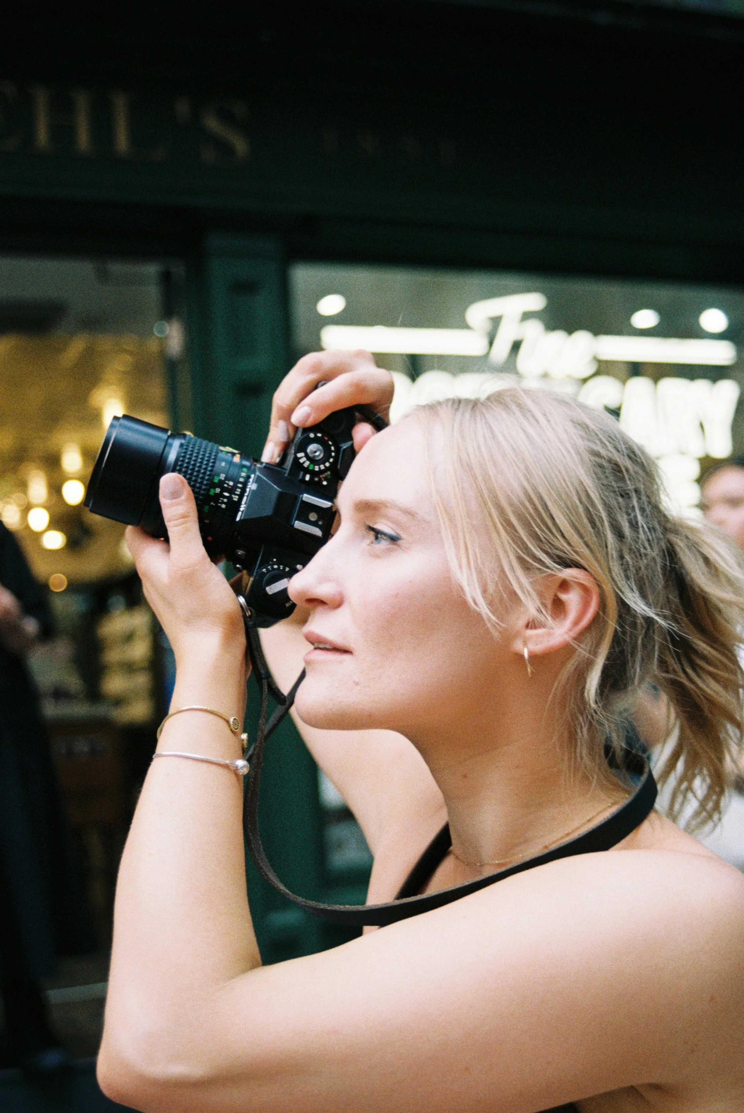 A young woman with blonde hair takes a photograph indoors, holding a camera up to her eye, with a hotel lobby or restaurant in the background.