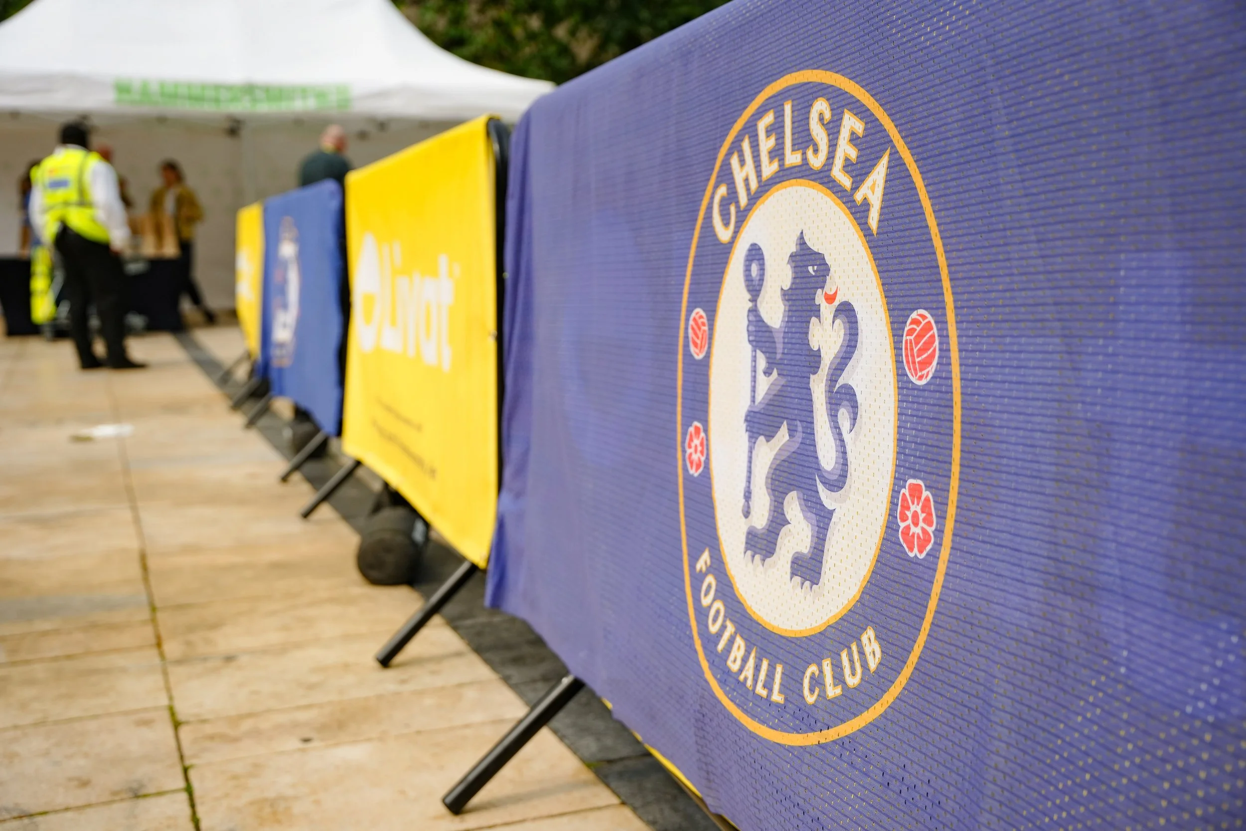 Chelsea Football Club logo on a blue banner at an outdoor event, with people and tents in the background.