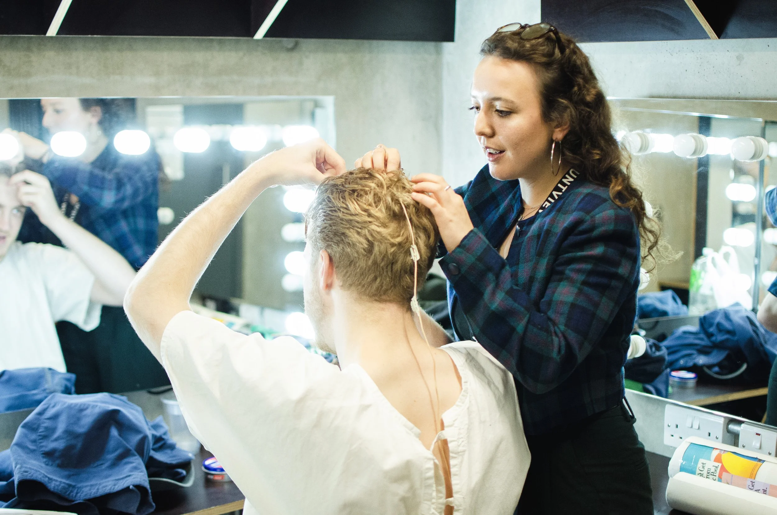 A makeup artist applying makeup to a male model with short, curly hair in a backstage dressing room. The room has bright mirror lights and various makeup and hair styling products on the counter.