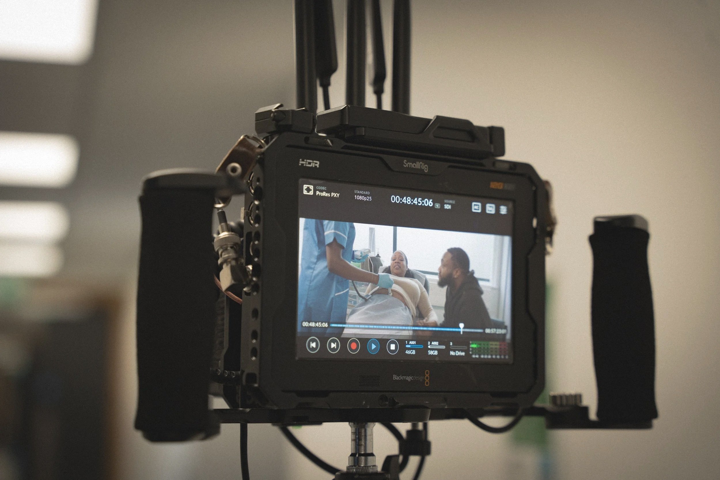 Video camera filming a scene with a woman lying in a hospital bed, attended by a nurse and a visitor in a hospital room.