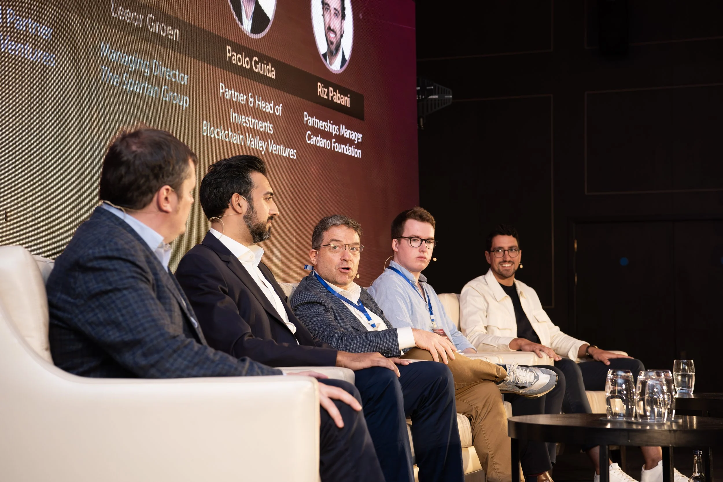 Panel of five men sitting on a stage in front of a large screen during a conference or panel discussion.
