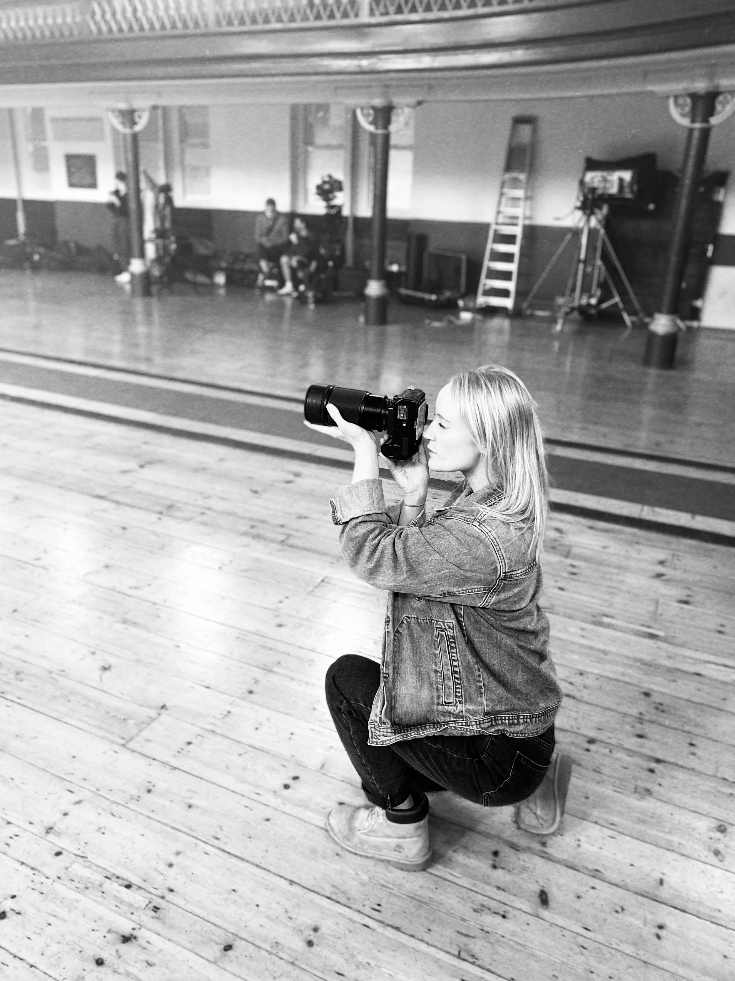 A woman crouching on a wooden floor, taking a photograph with a camera. She is wearing a denim jacket, dark pants, and sneakers. The background appears to be a studio or dance hall with a reflection in a mirror, a ladder, and two other people sitting and standing in the reflection.