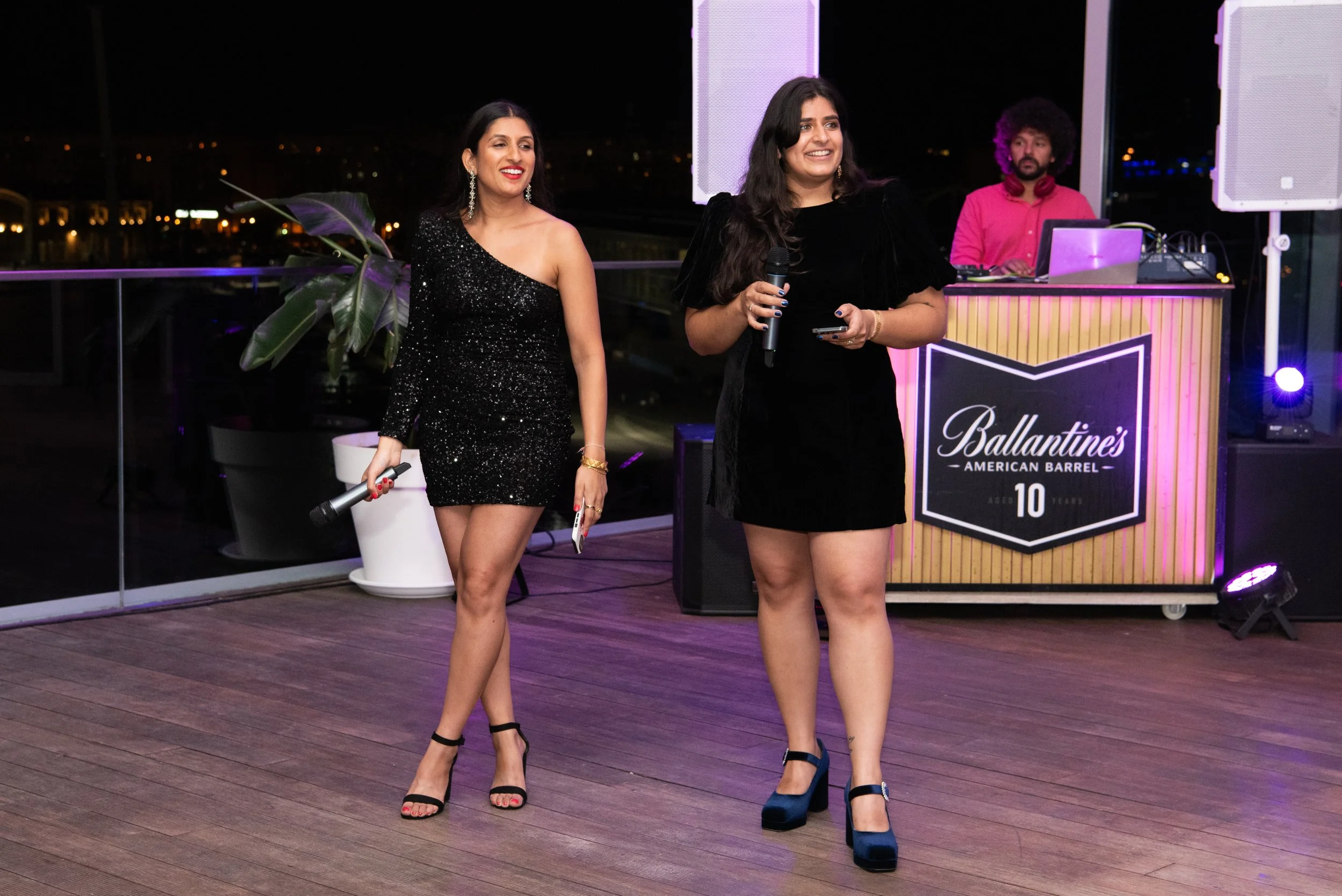Two women stand on a wooden floor with microphones in their hands, smiling at an event celebrating Ballantine's American Barrel's 10th anniversary. A DJ is in the background behind a stand with purple lighting.