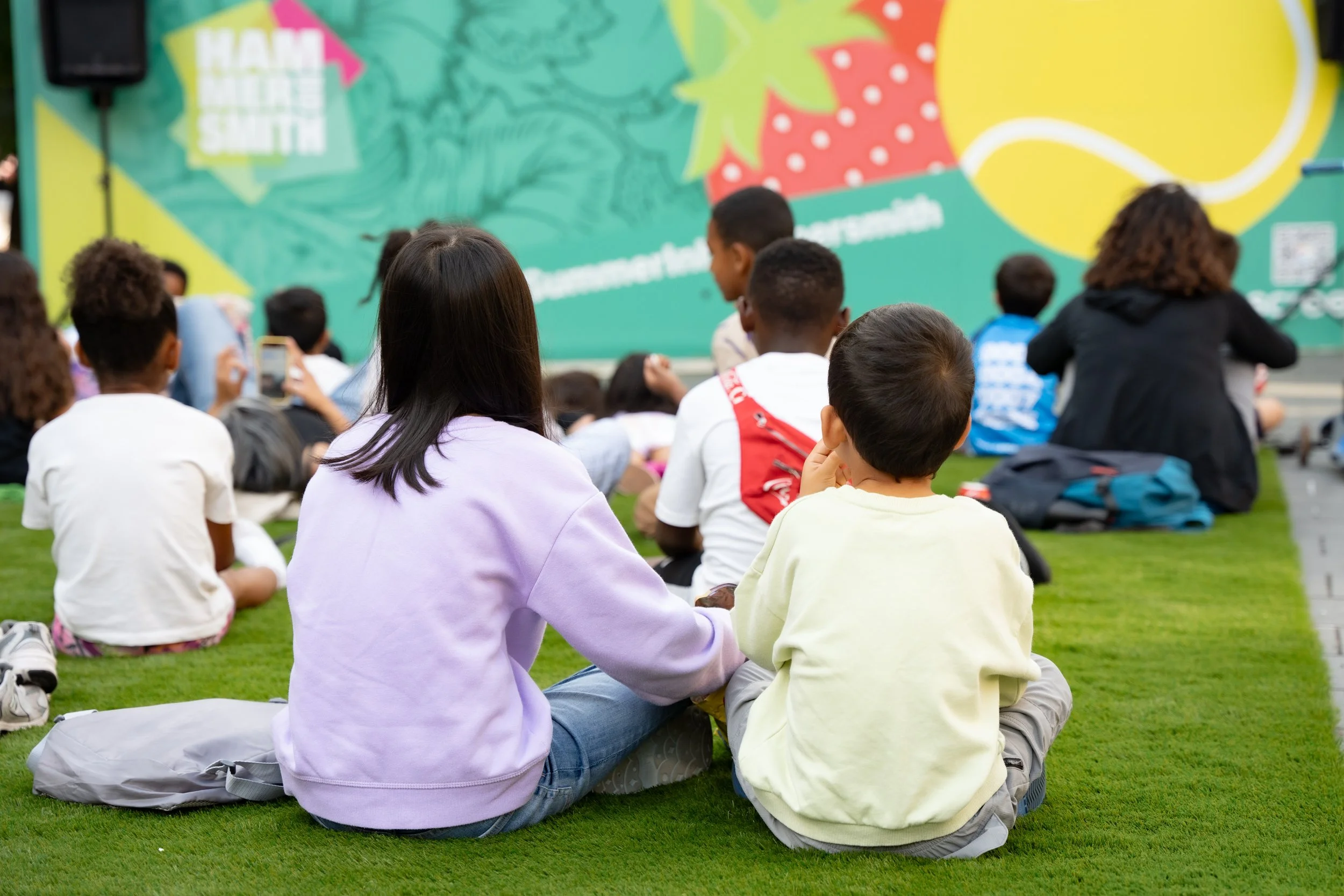 Children sitting on grass at an outdoor event, watching a presentation on a colorful stage with strawberries, lemon, and a green background.
