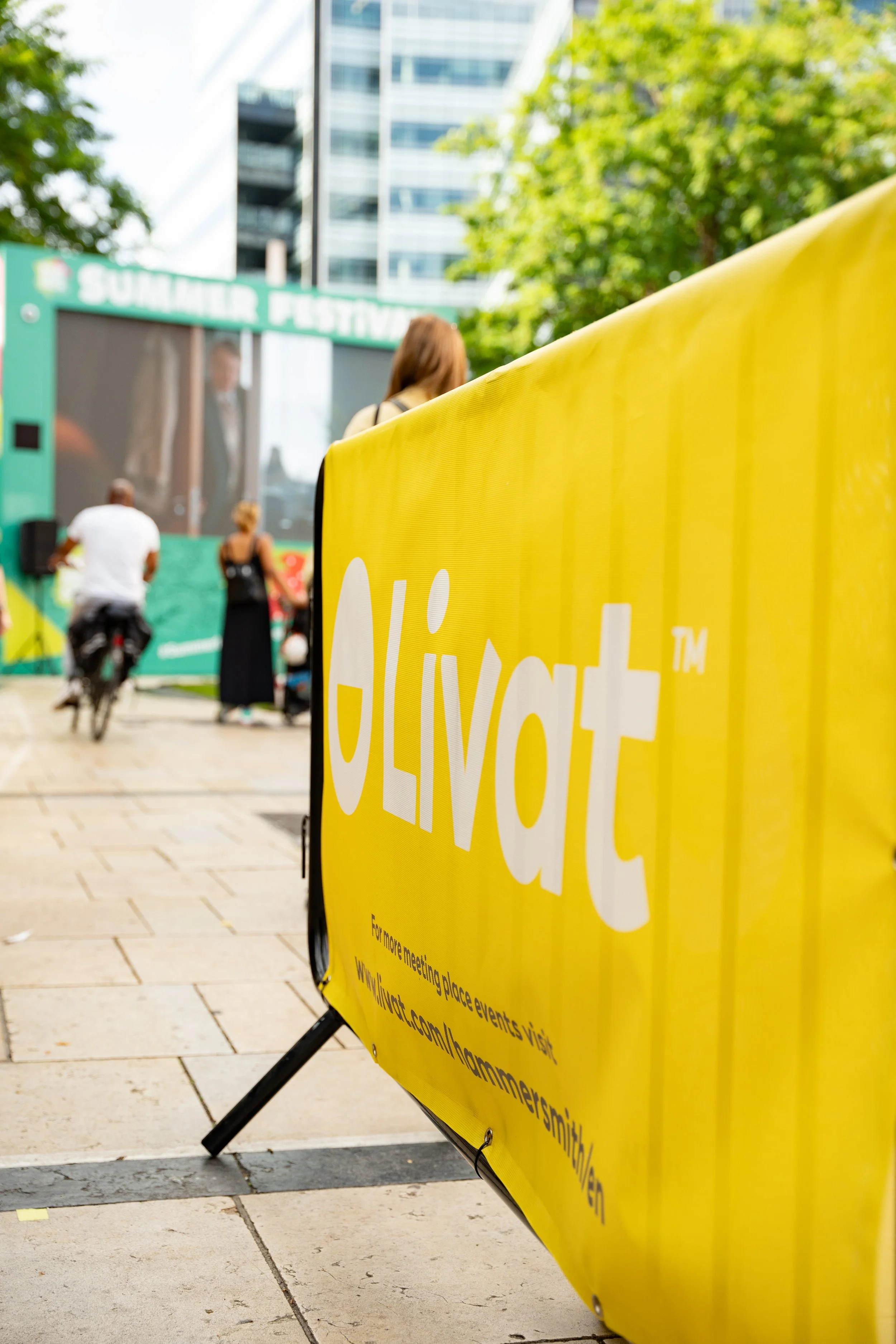 A yellow sign with the word 'Vote' in white letters, located outdoors at a summer festival with people, trees, and a large screen in the background.