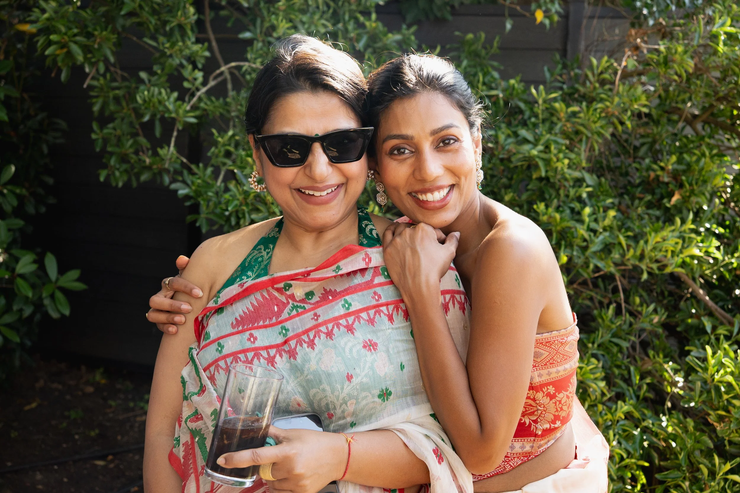 Two women smiling and hugging outdoors. One woman is wearing sunglasses, a colorful sari, and holding a glass of soda. The other woman is in a strapless dress, with her arm around the first woman, in front of green foliage.