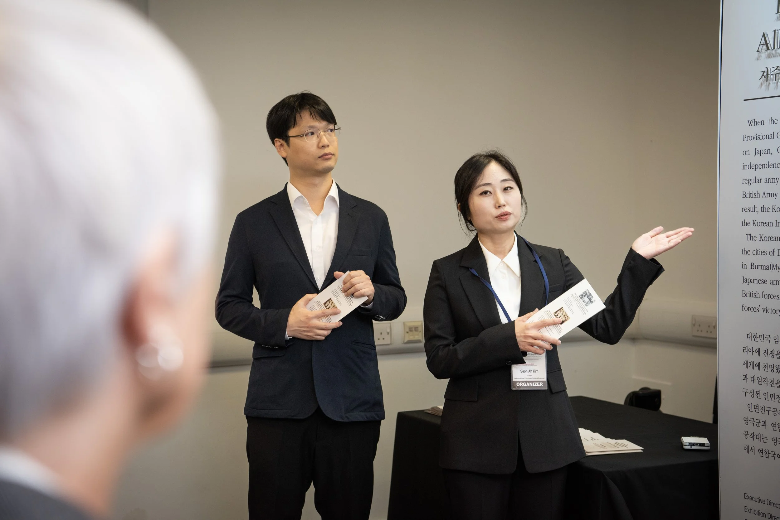 A woman in a black blazer and white shirt speaking and gesturing towards a large displayed document or presentation in a conference room, accompanied by a man in similar attire standing nearby.