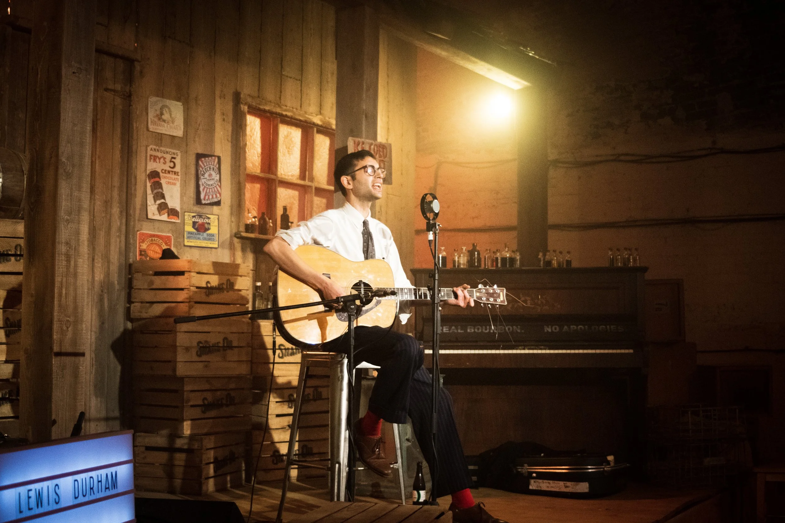 A man in a white shirt, tie, and glasses playing an acoustic guitar and singing into a microphone in a rustic wooden room with warm lighting, a piano, and vintage posters on the wall.