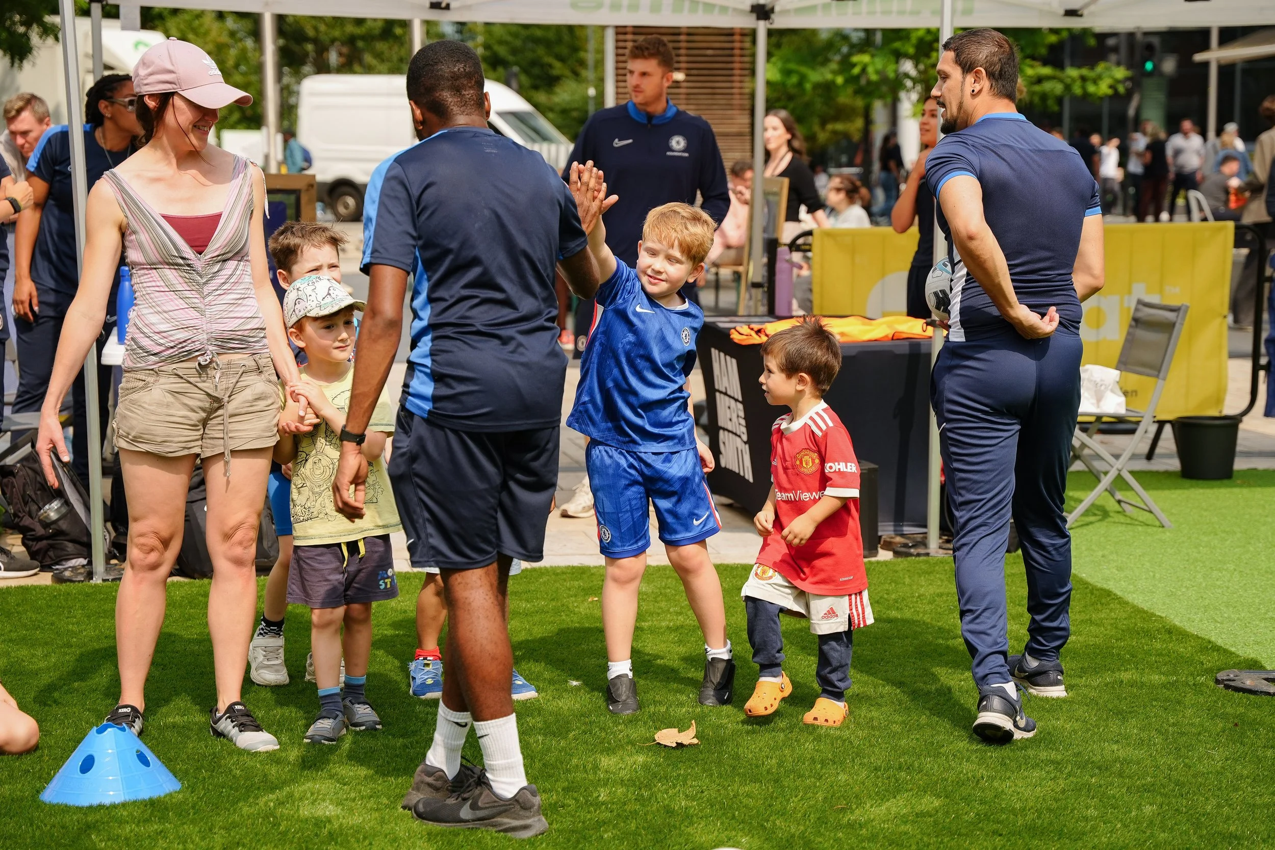 Children and adults on a grassy field, some wearing soccer jerseys, engaging in a high-five, with seating and people in the background.