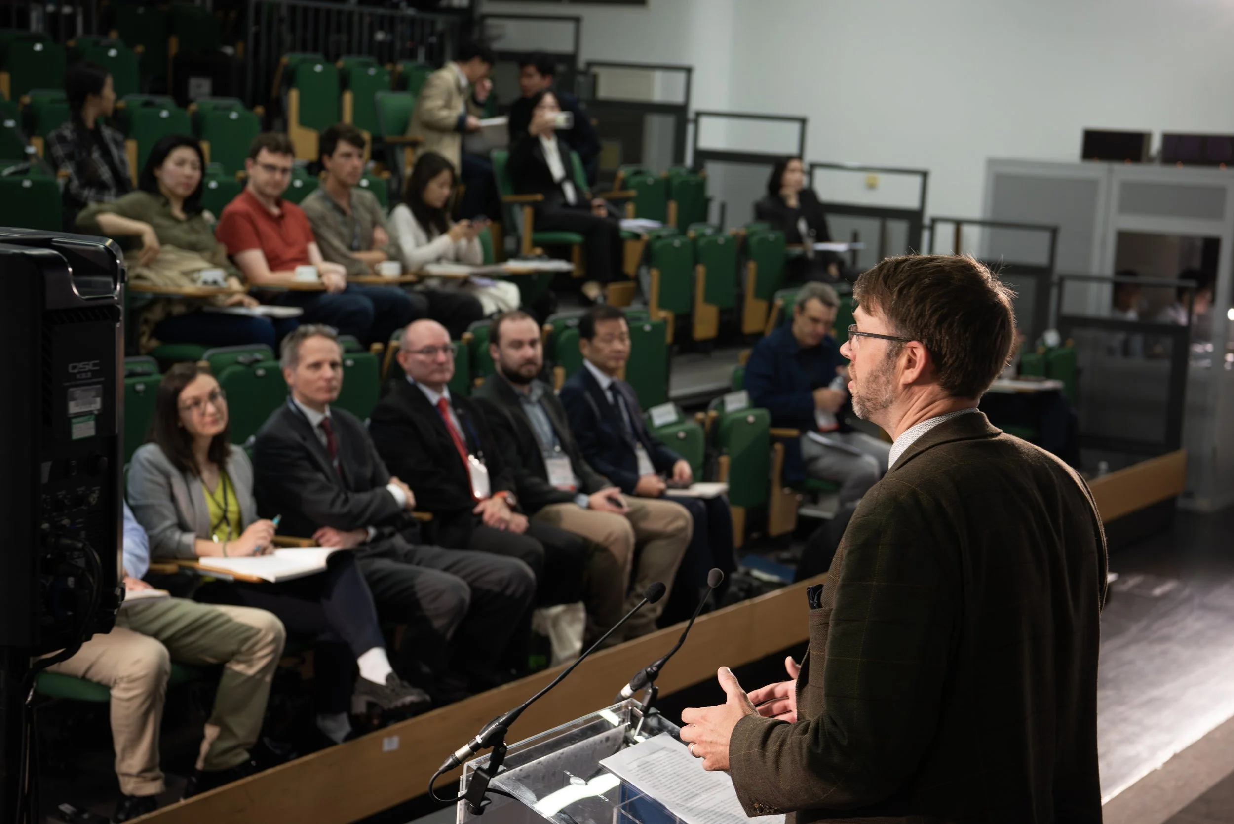 A man giving a presentation in a conference room with an audience of professionals. The audience is seated in green chairs, some taking notes, others listening attentively.